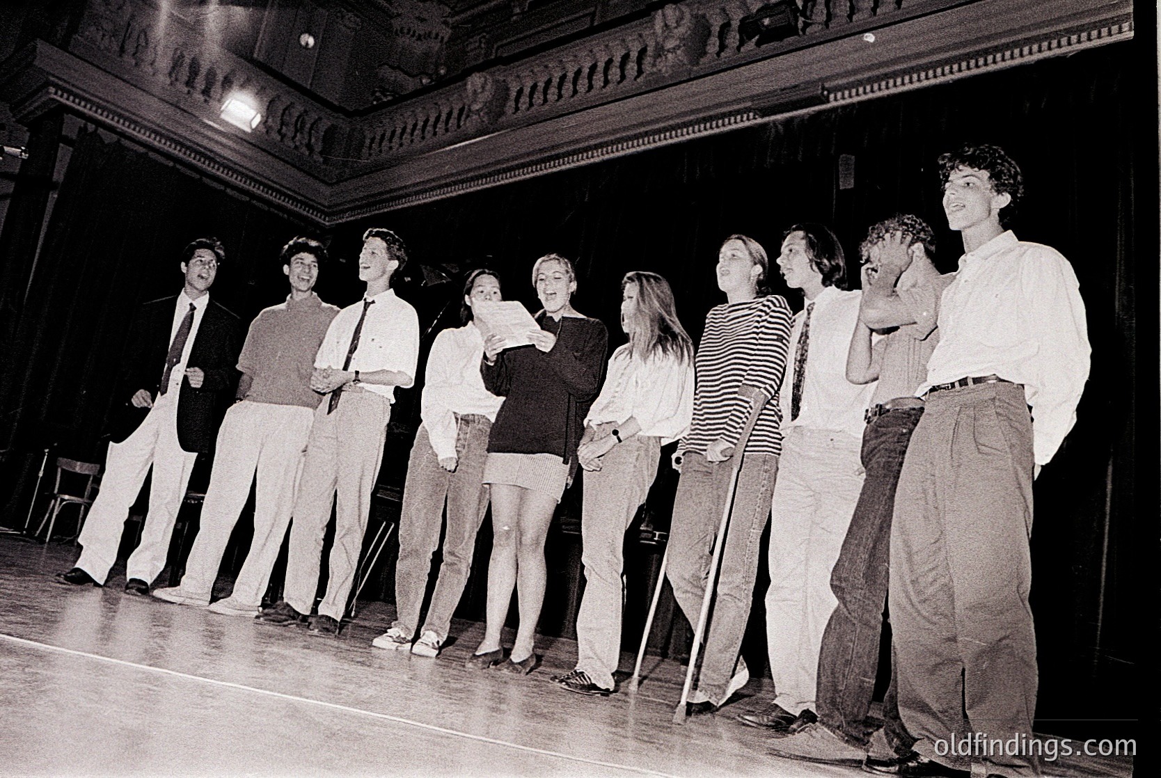 Black-and-white stage performance featuring 9 actors in 1970s attire—men in dress shirts/ties, women in blouses/skirts or pants—on a wooden stage with ornate ceiling details. Likely a theatrical production or rehearsal.