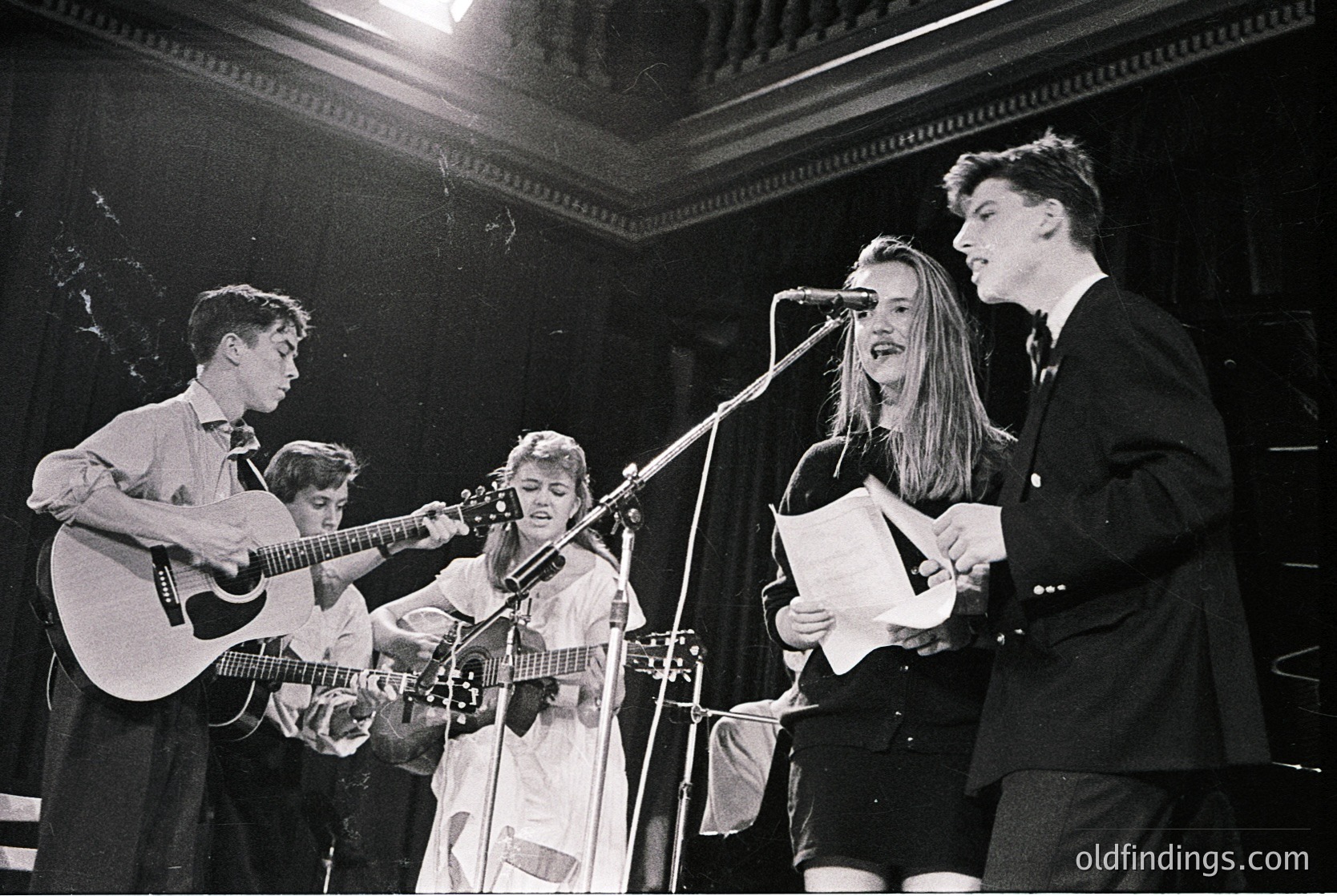 Vintage black-and-white photo of a folk/rock band performing live in a dimly lit indoor venue, likely 1960s–1970s. Four musicians: two playing acoustic guitars, one holding sheet music at a mic, and one singing into a microphone. Formal attire suggests a concert hall or cultural event. Authentic retro aesthetic for historical research or vintage design references.