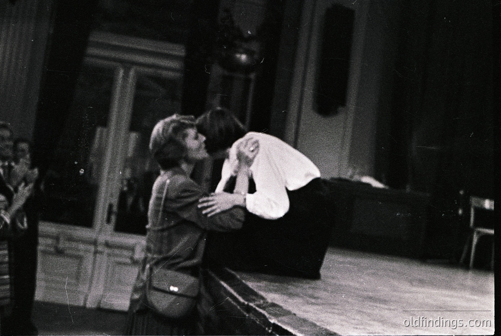 Two individuals embrace in an indoor setting, likely a theater or performance space. The person on the left wears a dark jacket with a visible pocket, while the other, in a light-colored dress, leans forward. Wooden stage elements and a hanging spotlight suggest a theatrical context. Style and lighting indicate a mid-20th century (1950s–1970s) production.