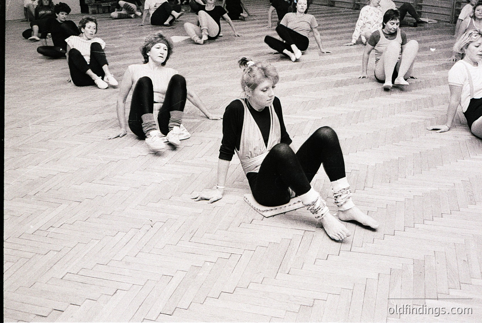 Group of women in mid-20th-century dance practice, likely 1960s–1970s, on polished wooden floor. Casual attire—leggings, sweaters, and socks—suggests informal rehearsal or fitness class. Indoor studio setting with natural light.