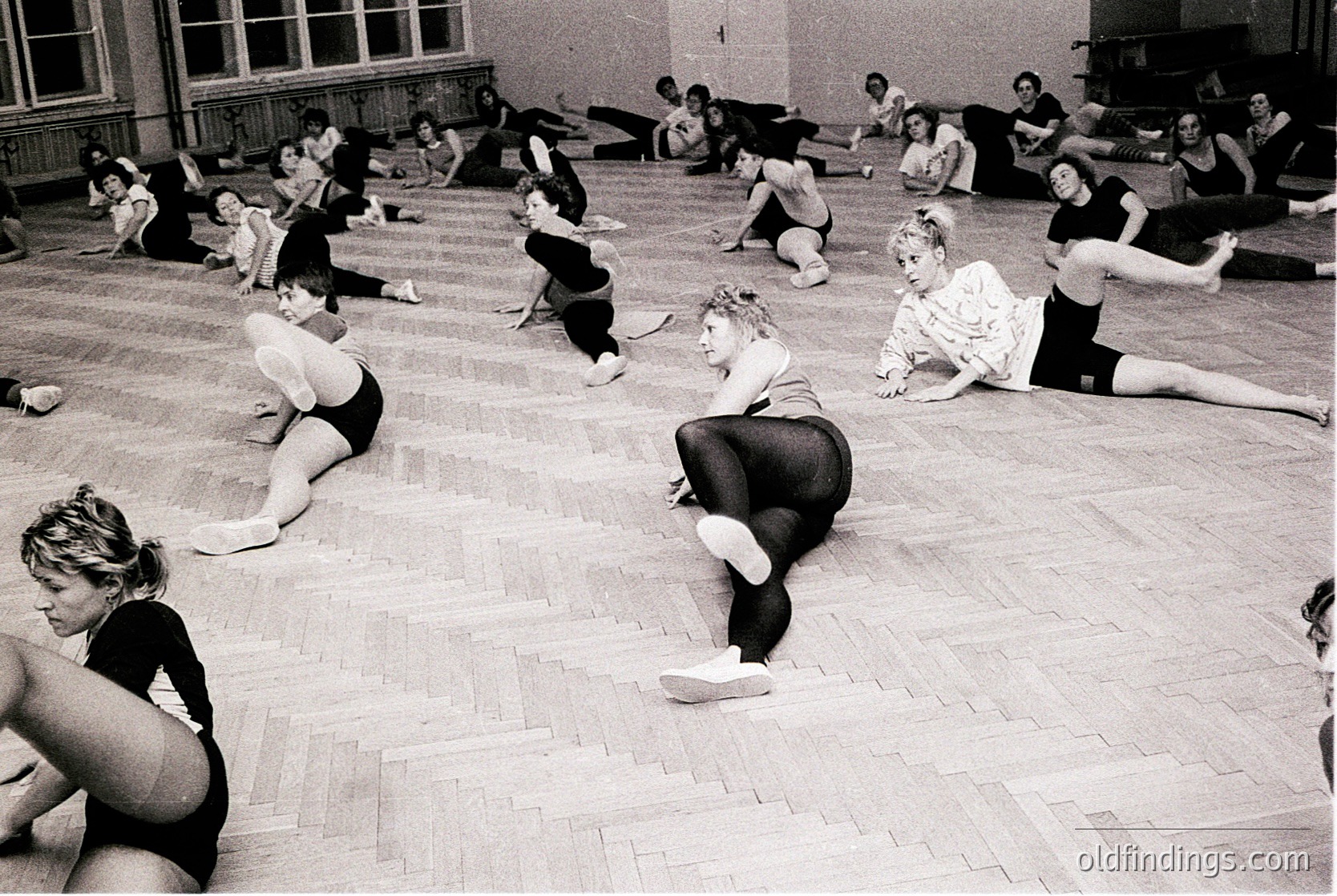 Vintage black-and-white photo of a ballet class in a large, wood-floored studio. Women in leotards and tights perform floor exercises, seated and kneeling in symmetrical lines. High ceilings, arched windows, and a fireplace frame the scene. Likely mid-20th century, Western Europe/US.