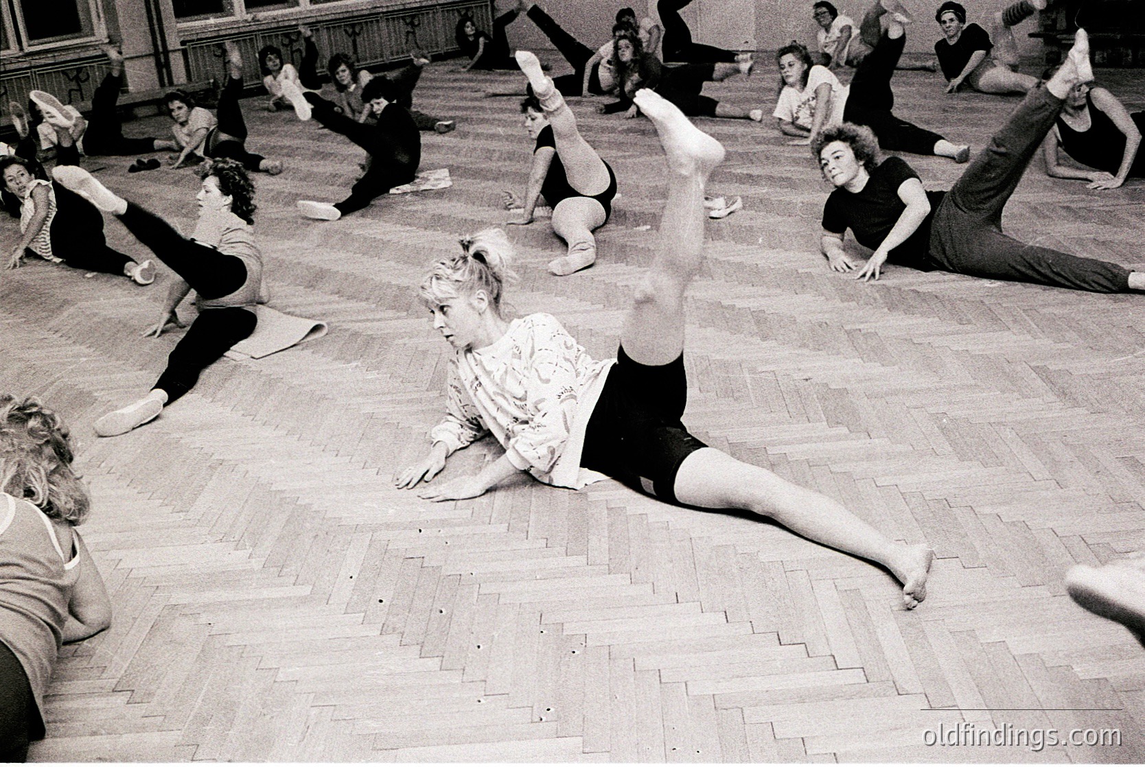Group fitness class in a spacious indoor gym, likely mid-20th century. Women perform floor exercises in coordinated poses, emphasizing leg stretches and balance. Wooden flooring and high ceilings suggest institutional or educational setting.