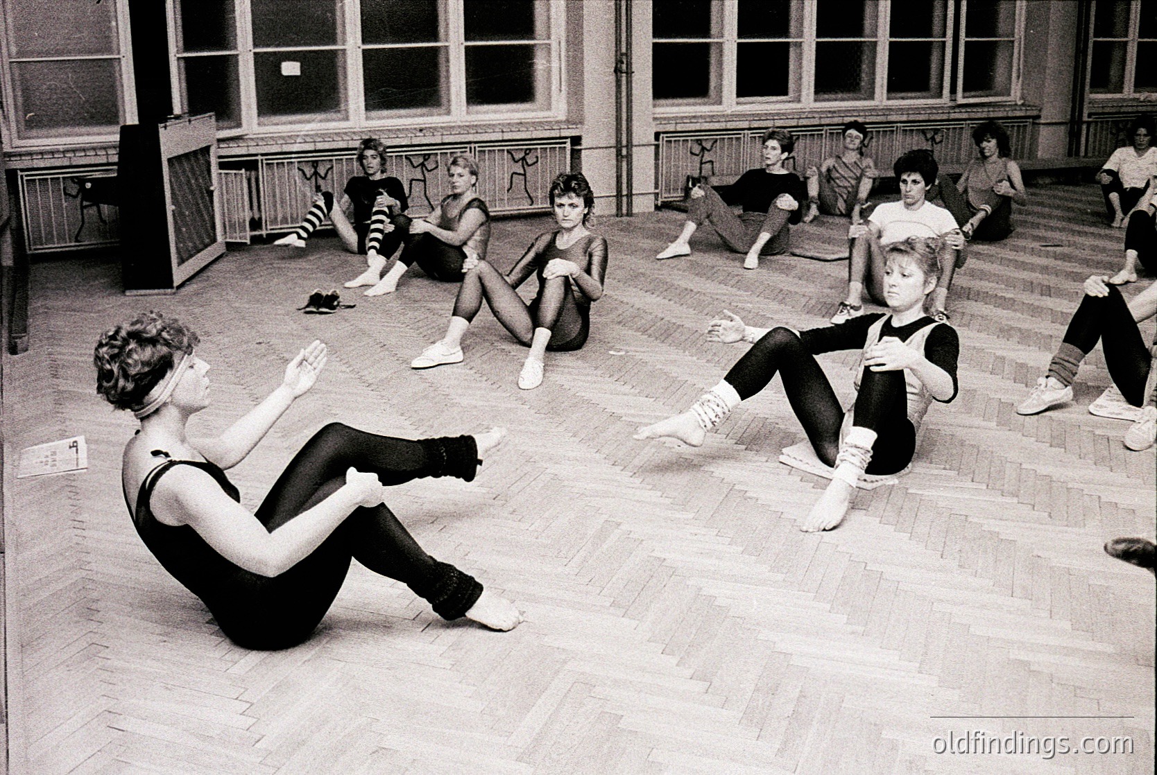 Black-and-white photo of a 1960s-70s ballet class in a large, wood-floored studio with high windows. Women in leotards and tights perform floor exercises, emphasizing form and alignment. Industrial-era architecture visible in background.