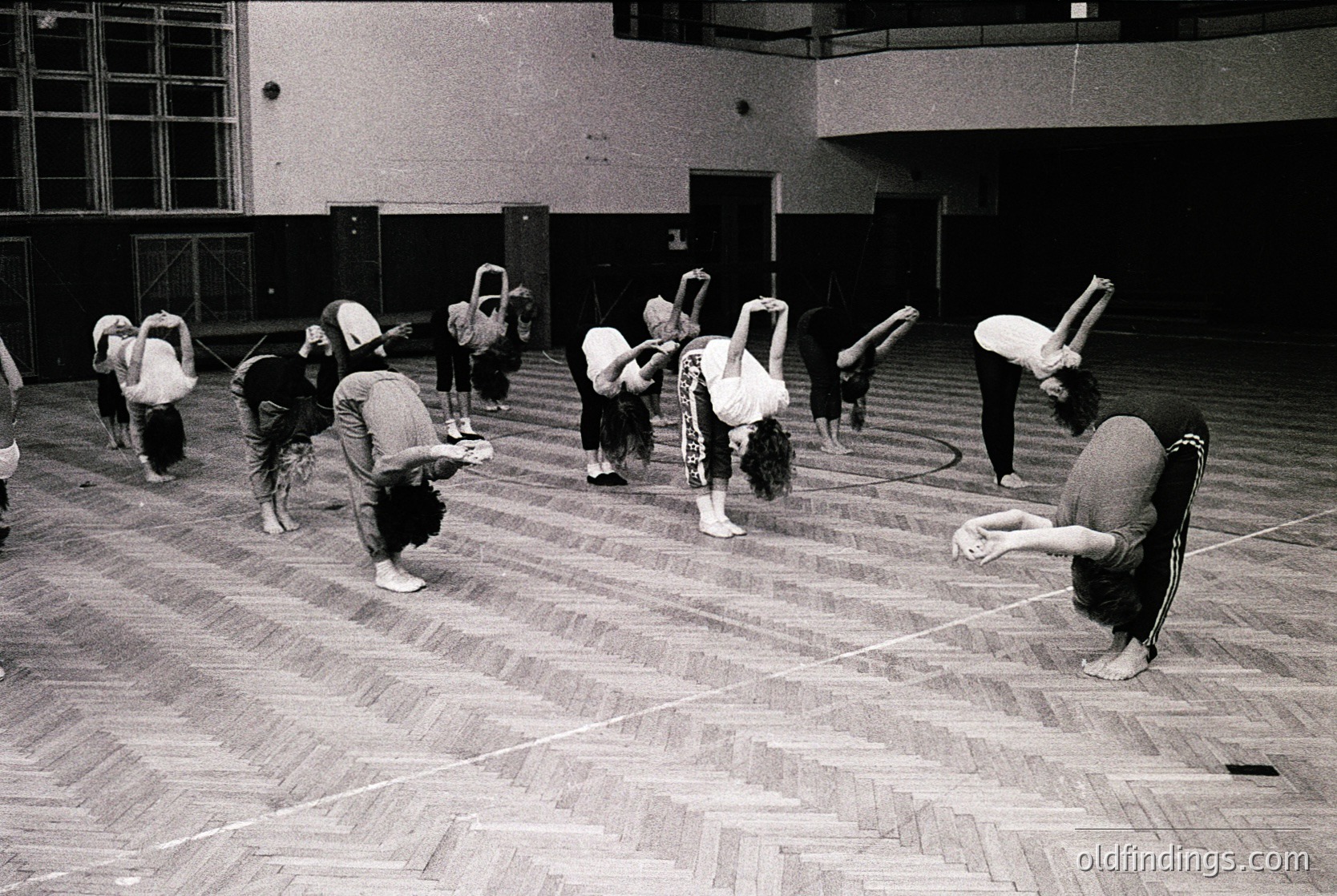 Group yoga/stretching session in an indoor gym, likely 1970s–1980s. Participants in loose clothing and headbands perform floor exercises on wooden parquet flooring. Minimalist gym architecture with concrete walls and large windows.