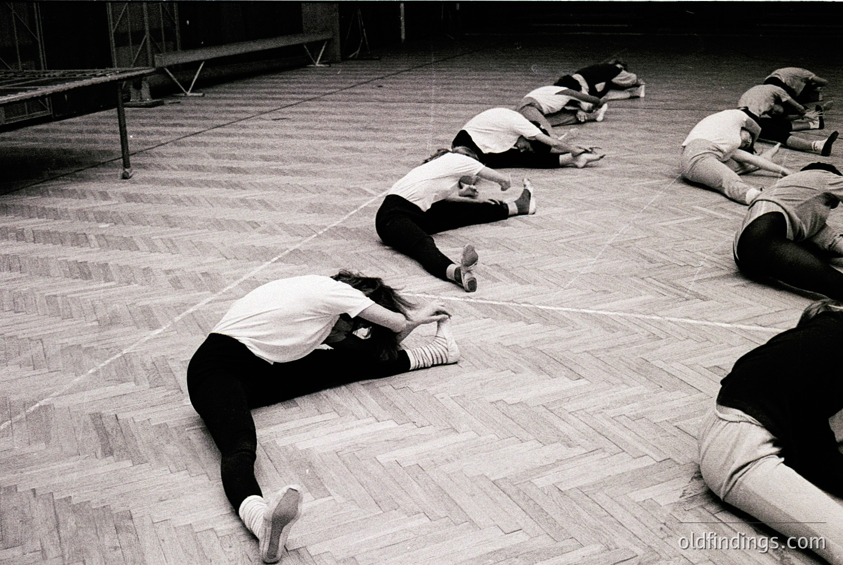 Group of individuals in mid-motion, likely practicing floor exercises or dance in a spacious indoor wooden-floored hall. Uniform attire suggests a structured class or rehearsal, possibly 1970s–1980s. Industrial lighting and minimal decor indicate a utilitarian or educational setting.
