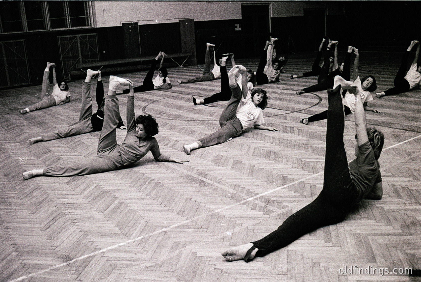 Group fitness class in a gymnasium, 1970s. Participants perform floor exercises in coordinated poses, likely inspired by Pilates or early yoga. Wooden floor and tiled walls suggest institutional or community center setting.