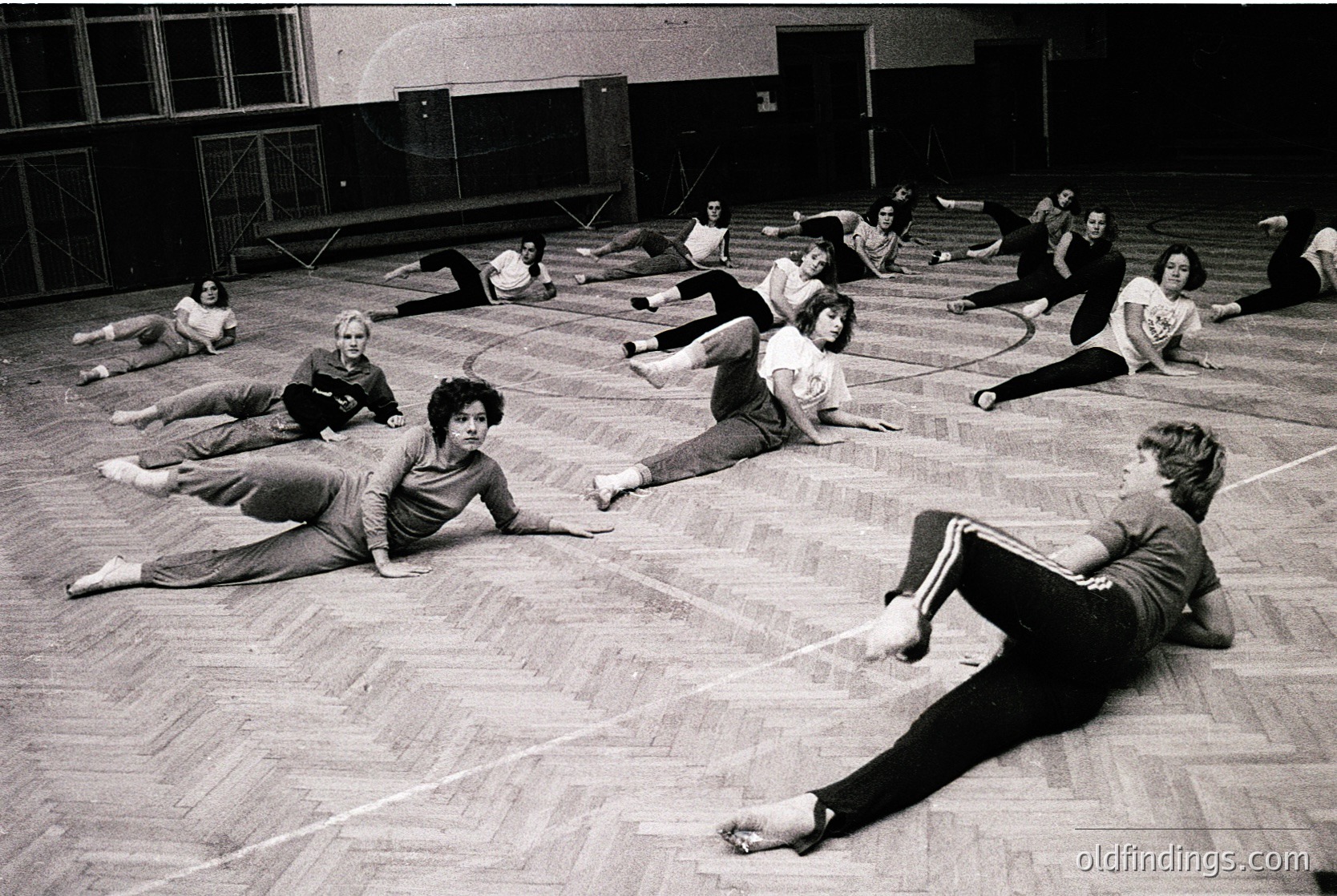 Group fitness class in a gymnasium, 1970s. Participants perform floor exercises in coordinated poses, likely part of a structured aerobics or movement program. Wooden floor and high ceiling with industrial lighting.