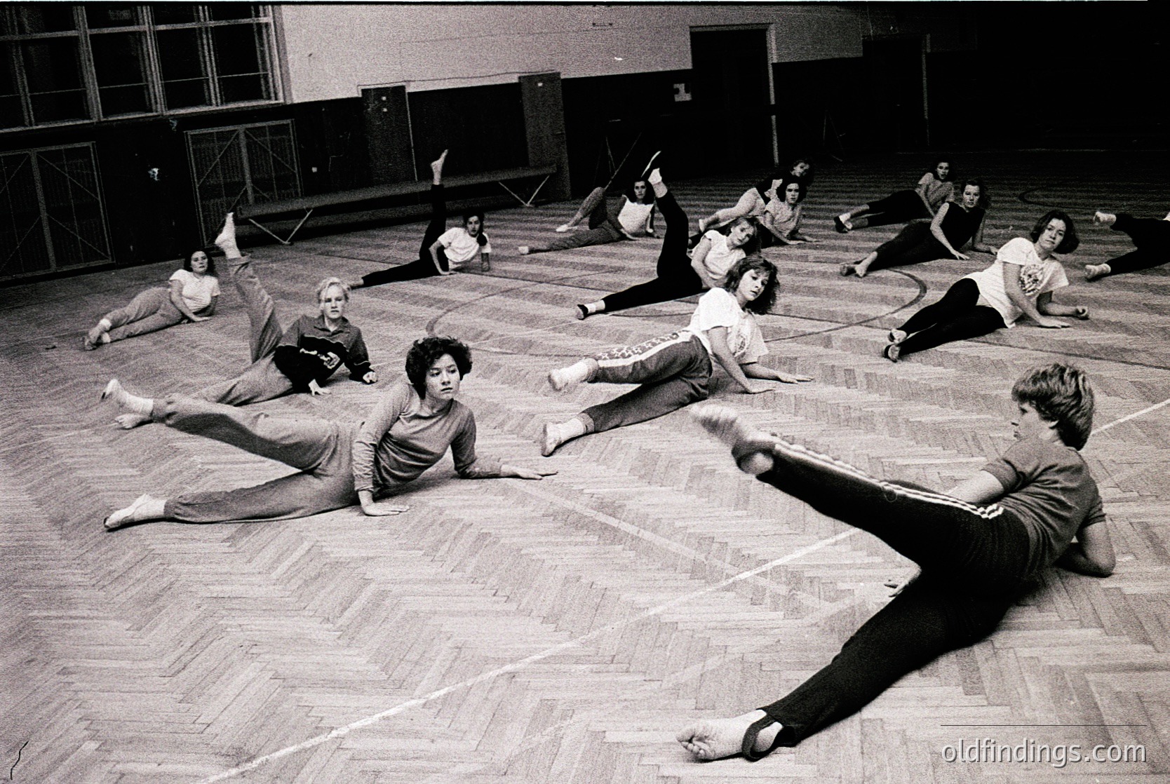 Black-and-white gym class in mid-20th century (likely 1960s–70s) with students performing floor exercises in a spacious indoor space. Hardwood floors, chalk lines, and a high ceiling with large windows suggest a school or community center setting. Dynamic poses reflect structured physical education routines.