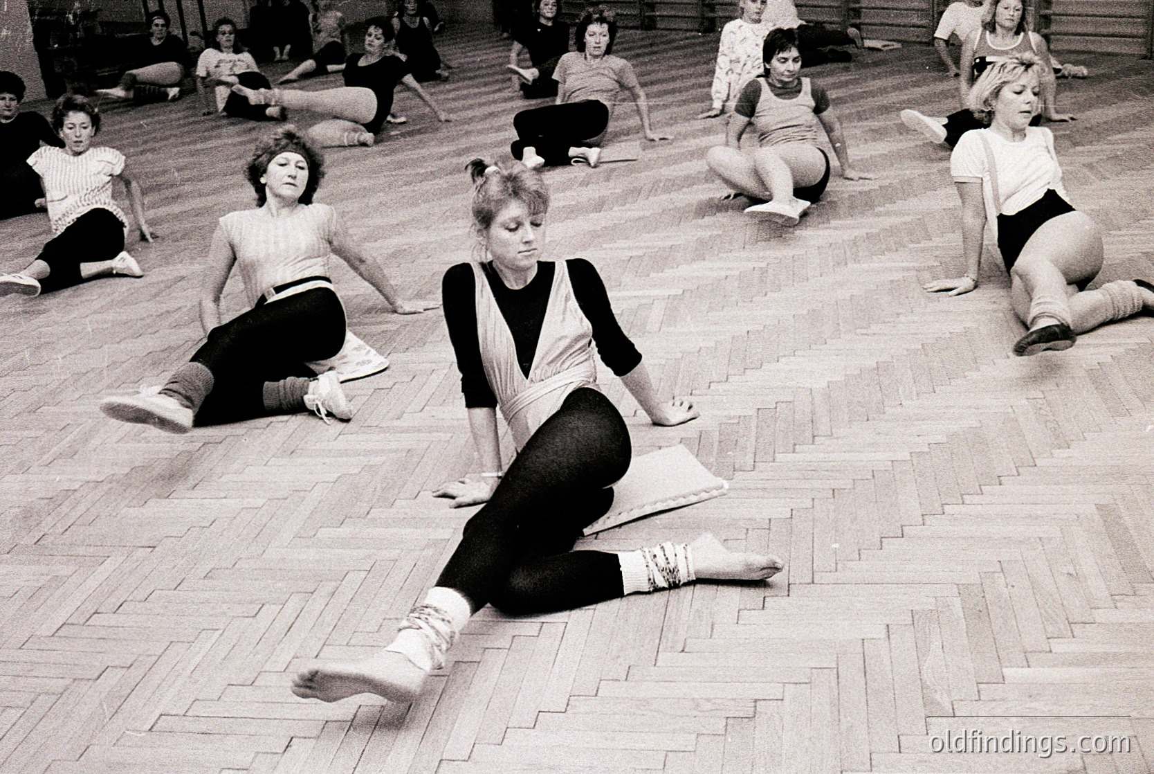 Group fitness class in a gymnasium, 1970s. Women in leotards and leg warmers stretch on wooden floors. Classic aerobic or yoga session, likely Eastern Bloc or Western Europe.