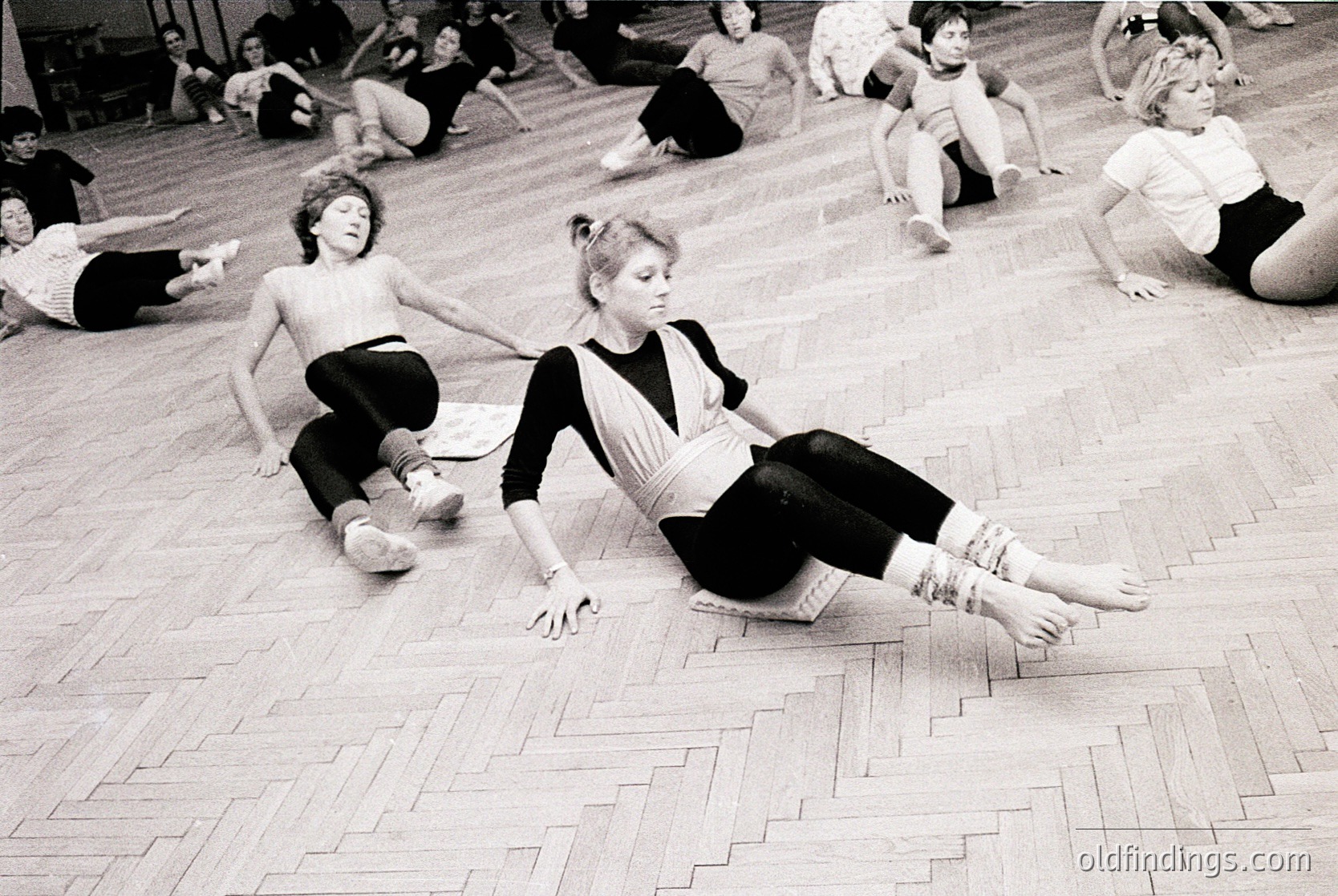 Group fitness class in a gymnasium, 1970s–1980s. Women in leotards and shorts perform floor exercises on wooden planks. Minimalist, indoor setting with natural light. Reflects early aerobics or dance fitness trends.