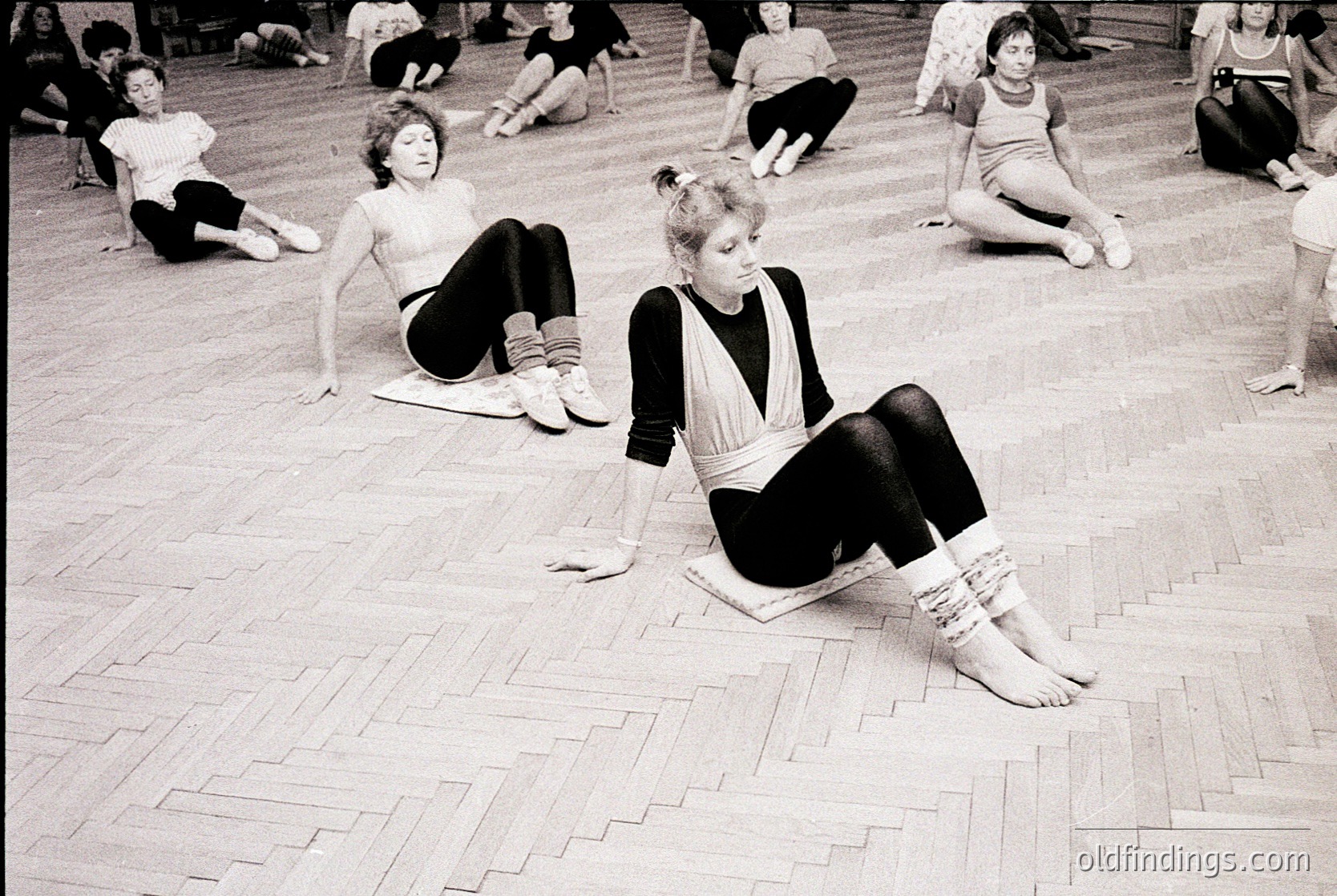 Classical ballet training session in a studio, mid-20th century. Dancers in leotards and tights practice floor exercises on polished wooden planks. Foreground dancer wears a black-and-white striped leotard and tights, seated in a split position.