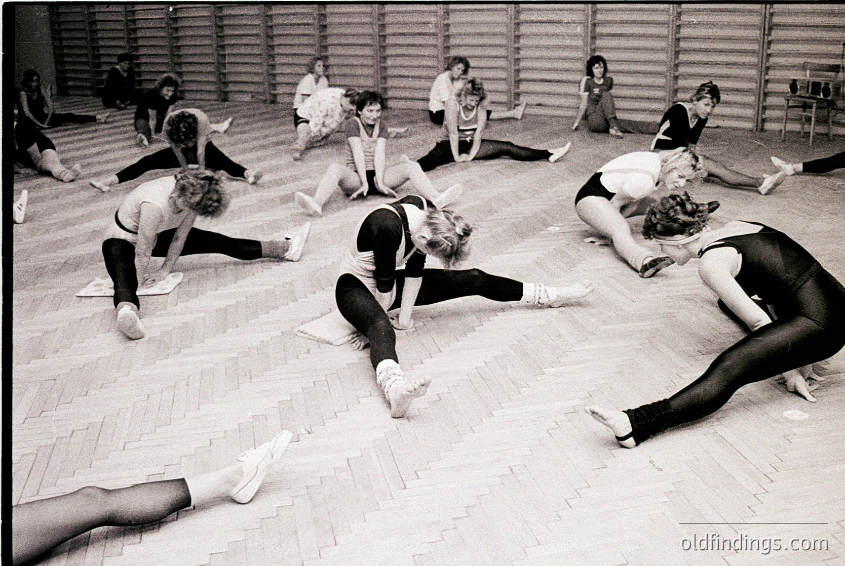Group ballet class in a wooden-floored studio, 1960s–1970s. Women in leotards and tights perform floor exercises, emphasizing flexibility. Minimalist composition highlights discipline and movement. Ideal for dance history, fitness, or vintage sports archives.