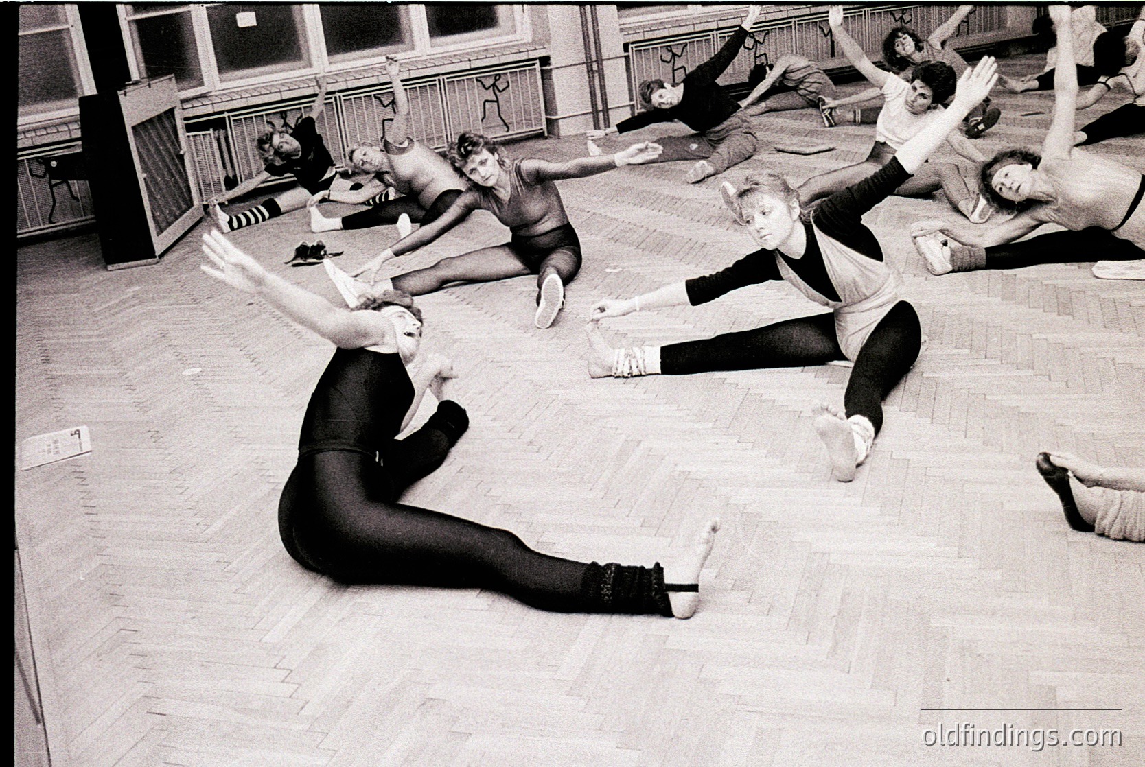 Group ballet class in mid-20th century studio, featuring dancers in leotards and tights performing floor exercises. High-arching wooden beams and vintage flooring suggest a mid-century ballet school.