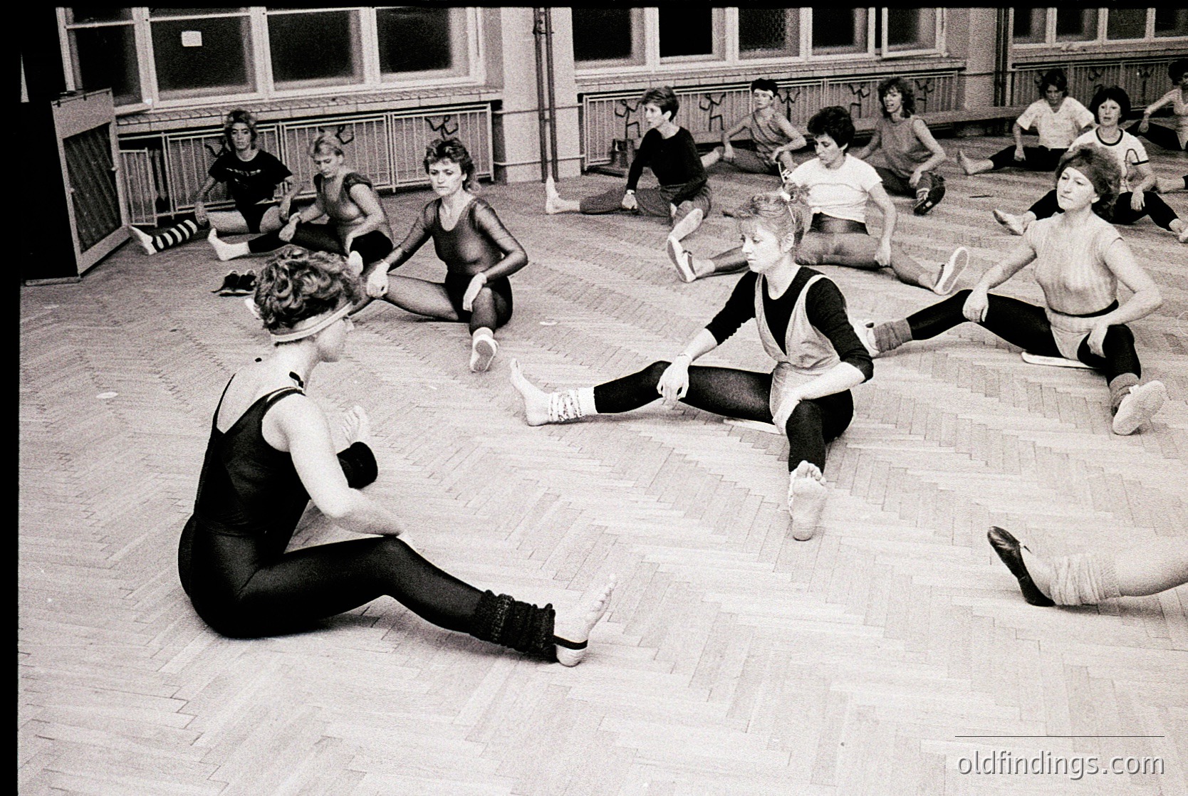 Black-and-white gym class featuring young students in leotards and tights performing floor exercises, 1960s–1970s. Wooden flooring and brick walls with windows suggest a school or community hall setting.