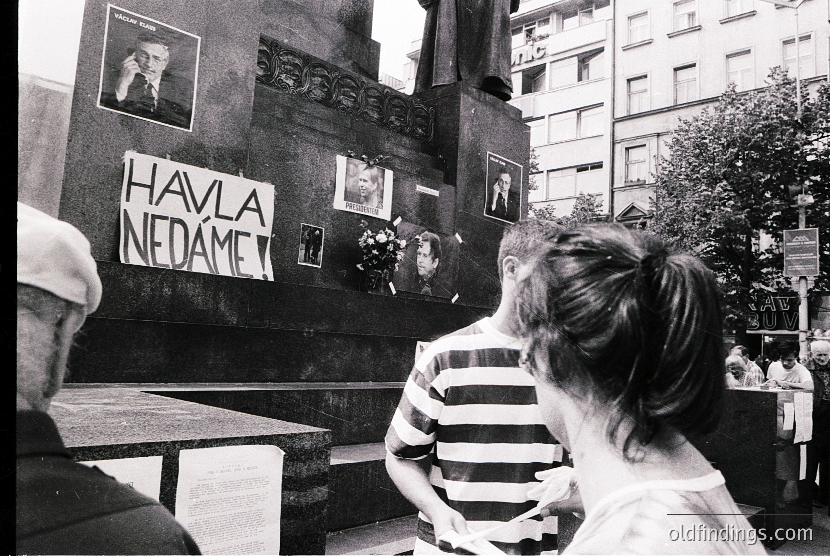 Black-and-white protest scene featuring a memorial wall with handwritten sign reading "HAVLA NEDÁME" (Czech/Slovak for "We Won’t Surrender"). Portraits and floral tributes attached to the wall. Urban setting with mid-rise buildings and blurred crowd in background. Likely 1980s Eastern Bloc, possibly Prague.