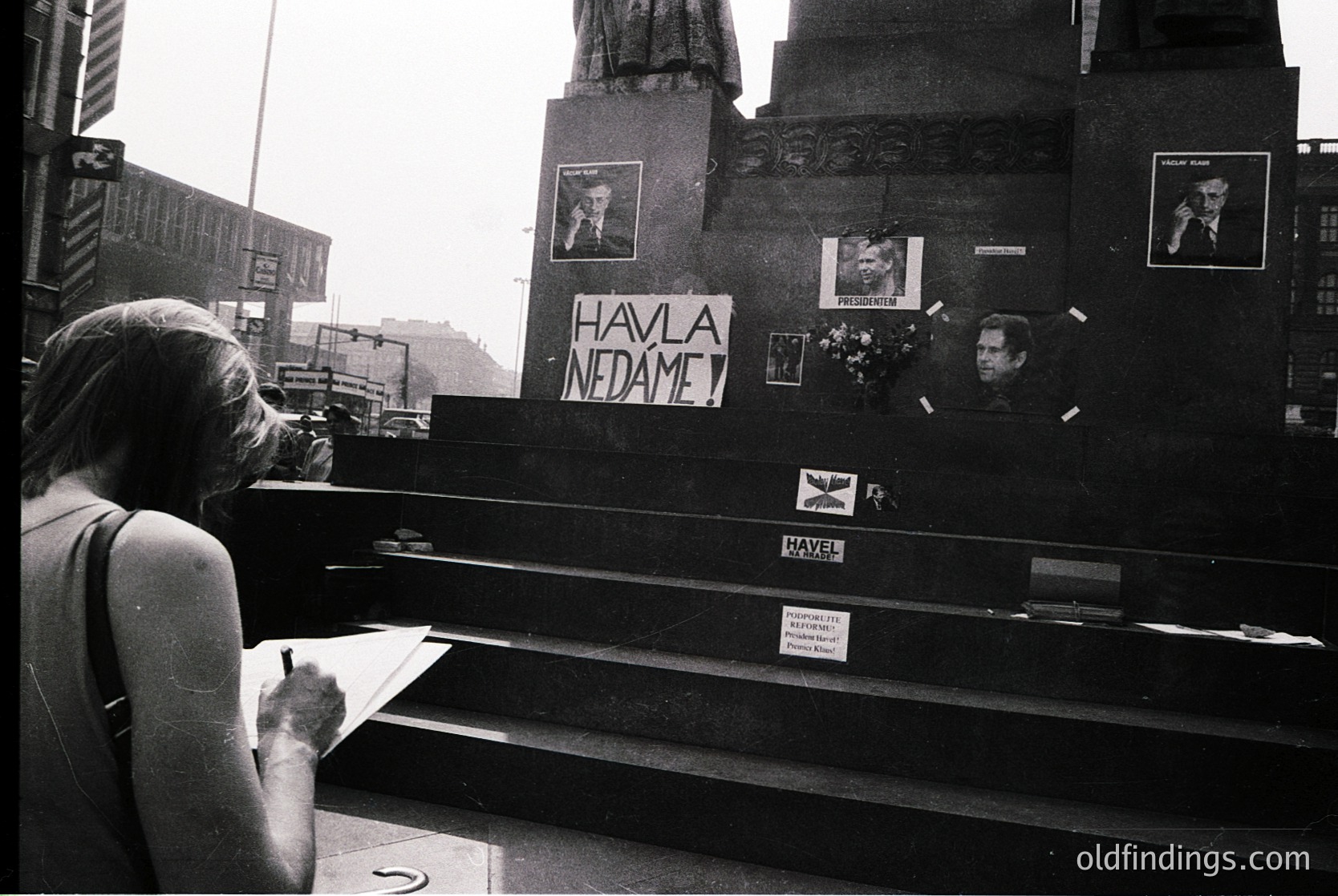 A woman in a sleeveless dress signs a memorial ledger at a public monument adorned with photos, flowers, and handwritten messages. Prominent sign reads *"Havla, nedáme"* (Czech: "Speak, we won’t forget"). Urban setting with Soviet-era architecture in background. Likely Prague, 1989 Velvet Revolution.