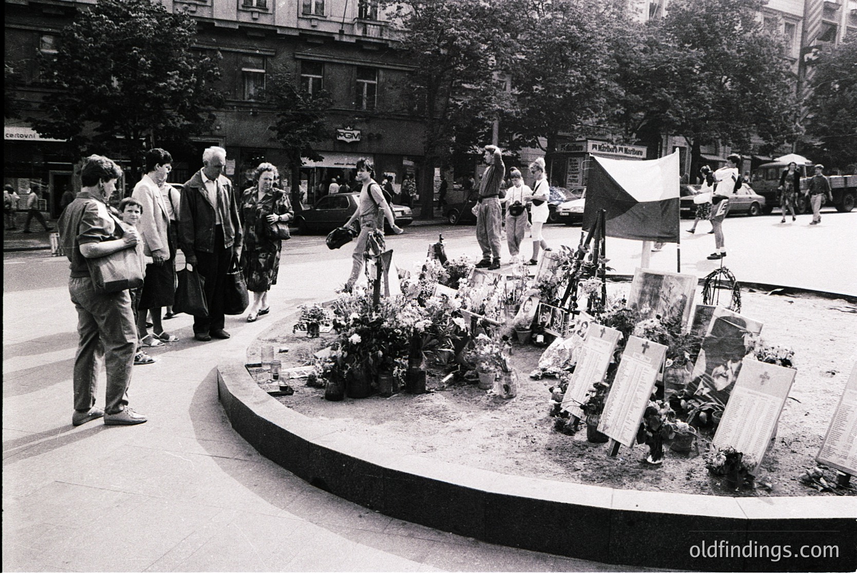 A solemn urban memorial scene from the late 20th century, likely Eastern Europe. A circular plaza features makeshift graves adorned with flowers, candles, and handwritten signs in Cyrillic script. A crowd of diverse ages stands in quiet reflection, some holding children. Surrounding buildings display Soviet-era architecture with signage in Bulgarian. The atmosphere suggests a public vigil or tribute.