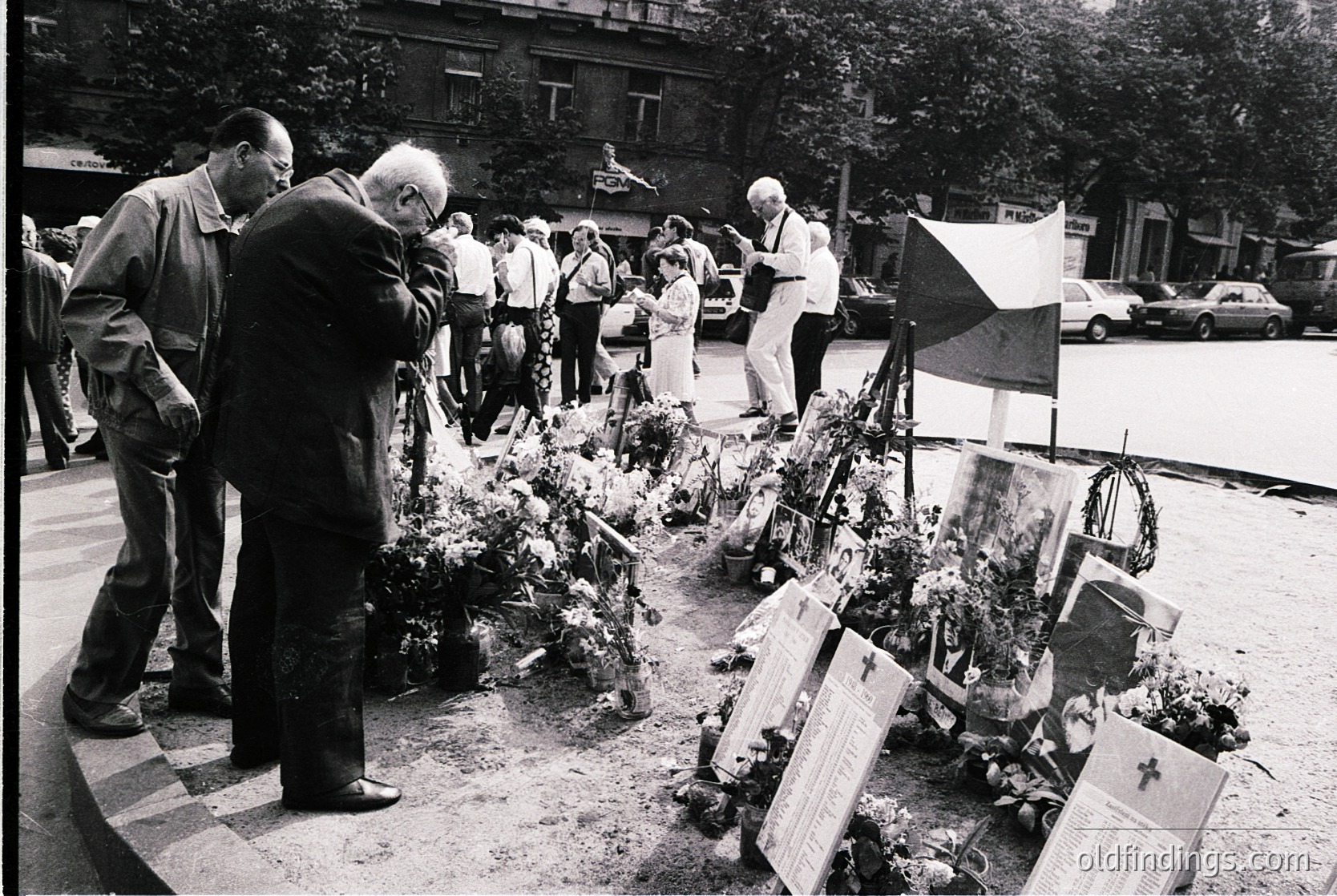 Black-and-white memorial scene with floral tributes and plaques arranged on a circular platform. Elderly men and women in 1970s-era clothing pay respects, some adjusting wreaths. Urban setting with Soviet-style architecture and parked cars in background. Likely a war or public tragedy memorial.