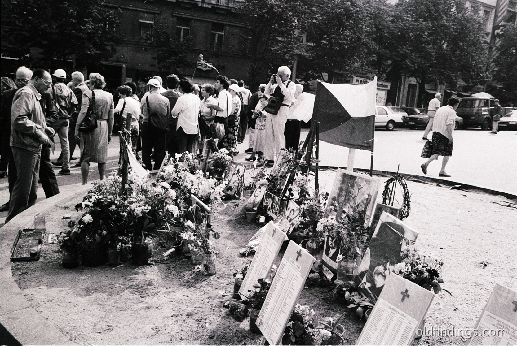 A solemn gathering at a memorial site, likely from the mid-20th century. Crowded urban street with people paying respects to a memorial adorned with floral tributes and plaques. The scene captures a moment of collective mourning, possibly post-war or commemorative. Urban architecture and vintage vehicles suggest a European city setting.