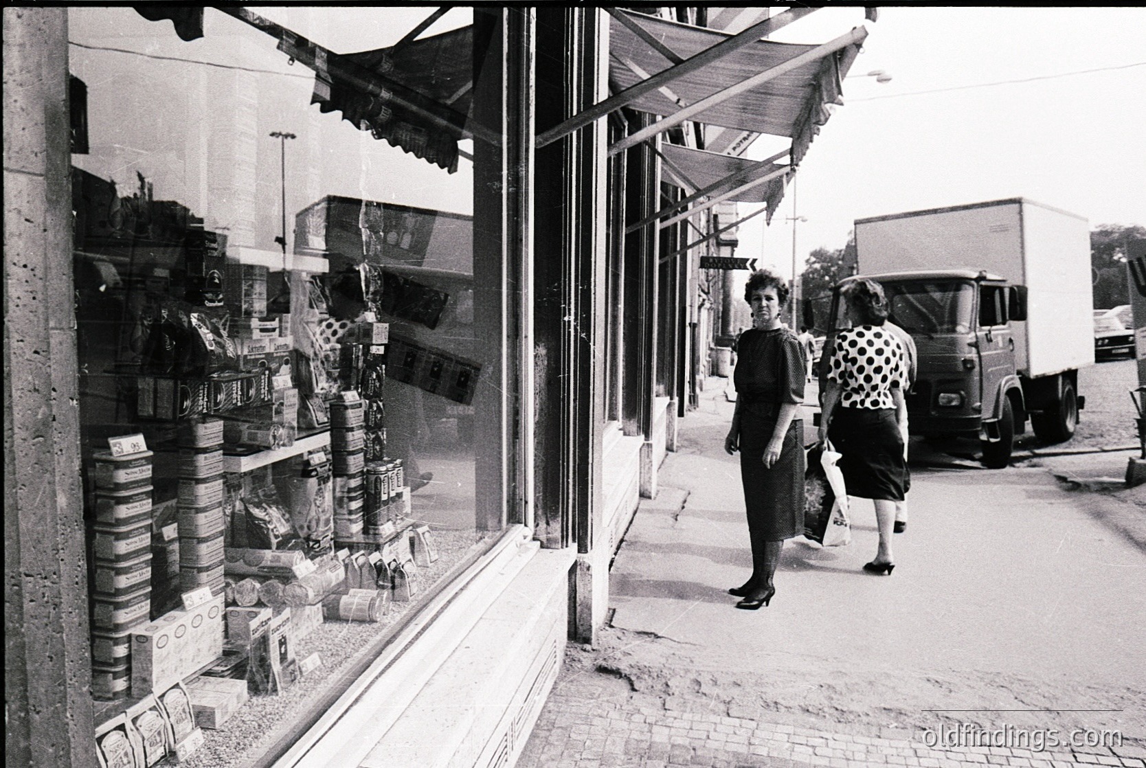 Vintage black-and-white street scene featuring a corner grocery store with shelves stocked in stacked cardboard boxes and jars. Two women in 1960s-era dresses walk past a parked delivery truck. Urban setting with cobblestone pavement and awnings.