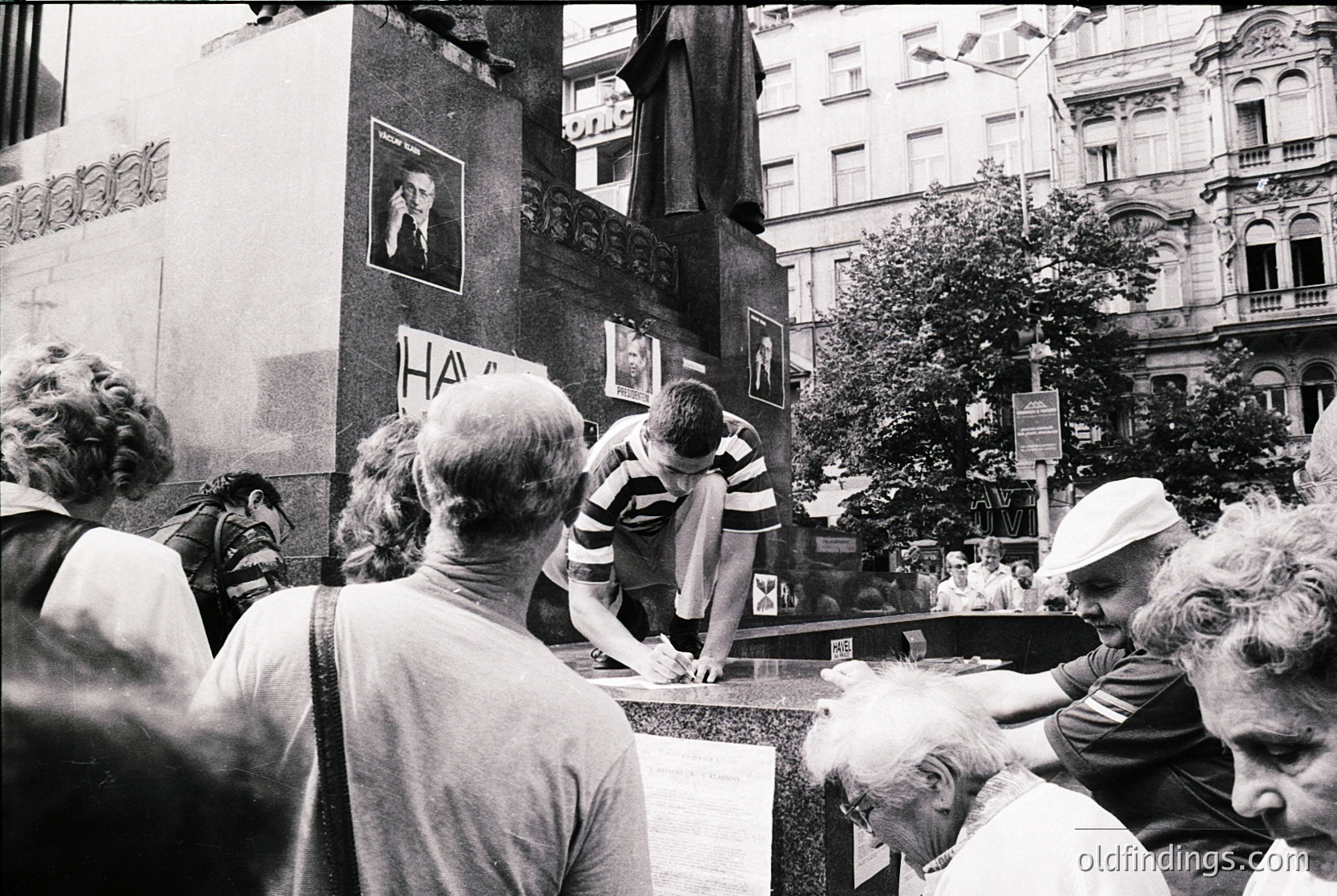 A candid 1970s street scene in a European city, likely . A group of people gathers around a makeshift barricade, with one man in a striped shirt leaning over a low wall. Posters and graffiti adorn the ornate building facade, hinting at political or cultural unrest. Urban architecture and vintage fashion capture the era’s social dynamics.