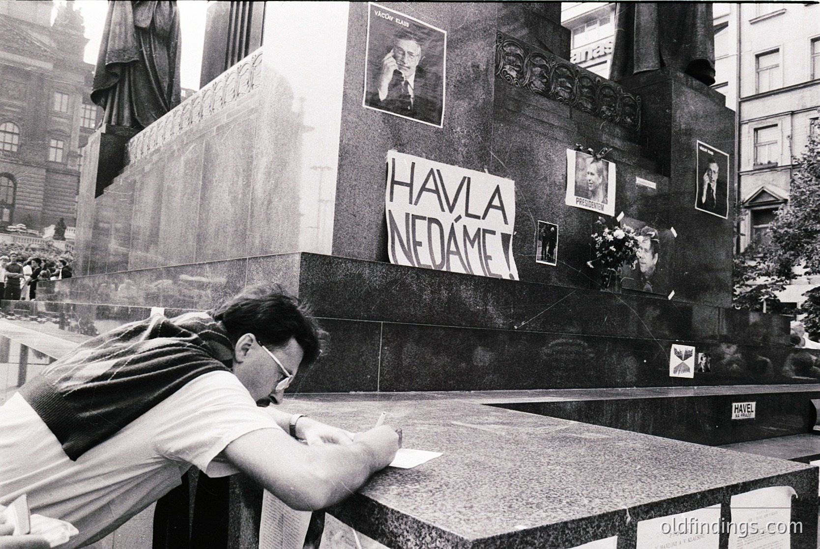 A man in a urban setting signs a memorial wall with the slogan "Havláč nedáme" (We Won’t Give Up Havlíček) in Prague, likely during the . Surrounding photos depict historical figures, and a statue of a seated figure looms in the background.