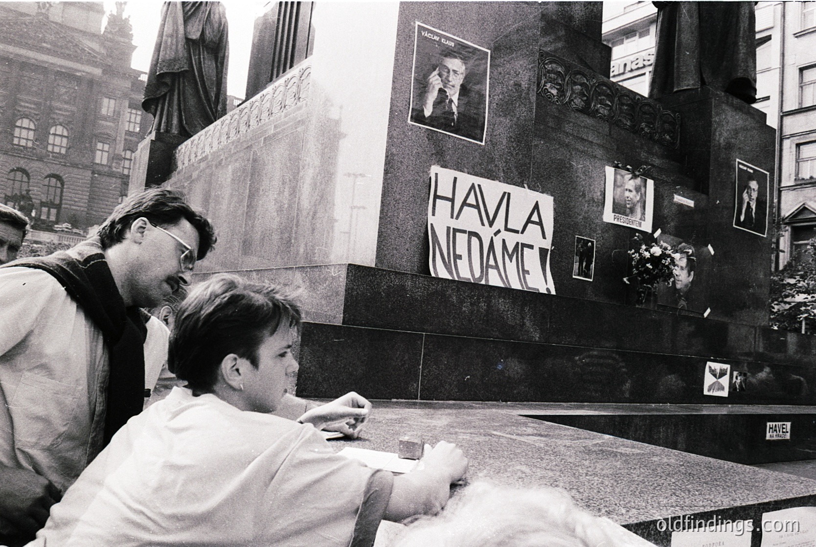 Two individuals examine graffiti reading *"Havlá nedáme"* (We won’t give up Havel) on a monument in Prague’s Wenceslas Square, 1989. Black-and-white protest imagery captures the Velvet Revolution’s public defiance.