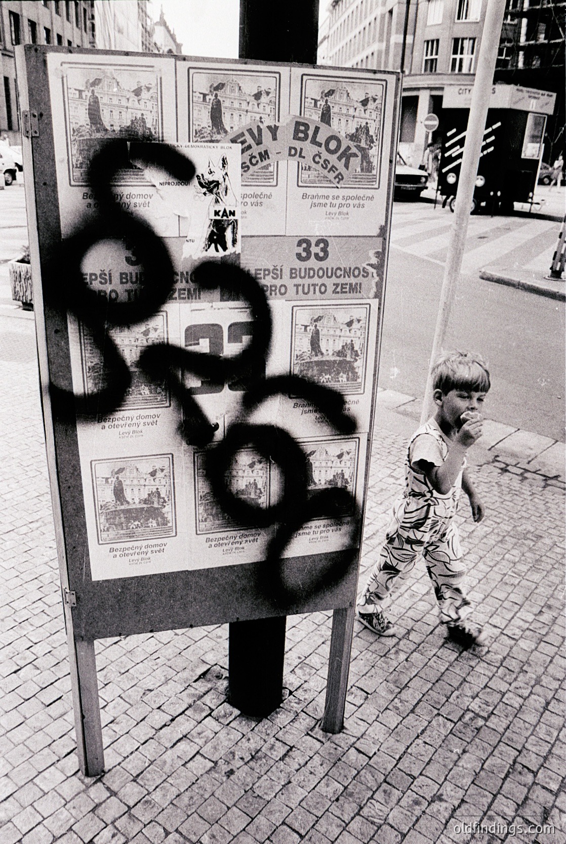 Vintage urban poster board covered in graffiti, featuring Cyrillic text and illustrations of figures in traditional attire. A child in shorts stands nearby, observing. Likely Eastern European, mid-20th century.