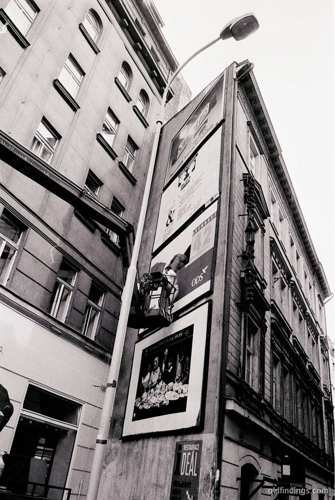 Mid-century urban facade featuring vintage billboards on a multi-story building. Prominent ads include a 1960s-era perfume campaign and a 1970s-style shoe advertisement. Architectural details include tall, narrow windows and ornate stonework. Likely European city street, possibly or .