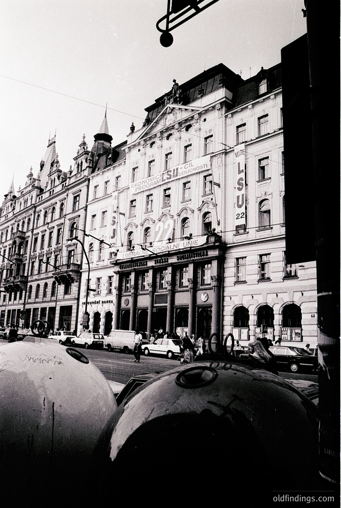 Historic European street corner with ornate 19th-century architecture. Prominent corner building features intricate stonework, arched windows, and a clock. Signage includes "SU" (likely a hotel) and "Savoy" hotel. Vintage cars and pedestrians suggest mid-20th century urban life.