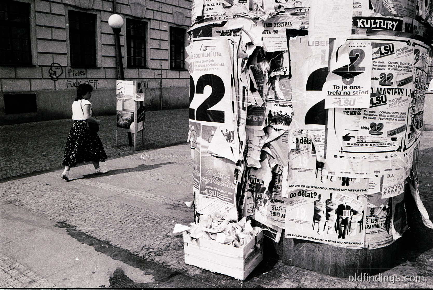 Vintage black-and-white street scene featuring a covered post office box overflowing with stacked flyers and ads. A woman in a patterned dress walks past, carrying a bag. Urban architecture with graffiti on walls and a lamppost in the background. Likely Eastern European, mid-20th century.