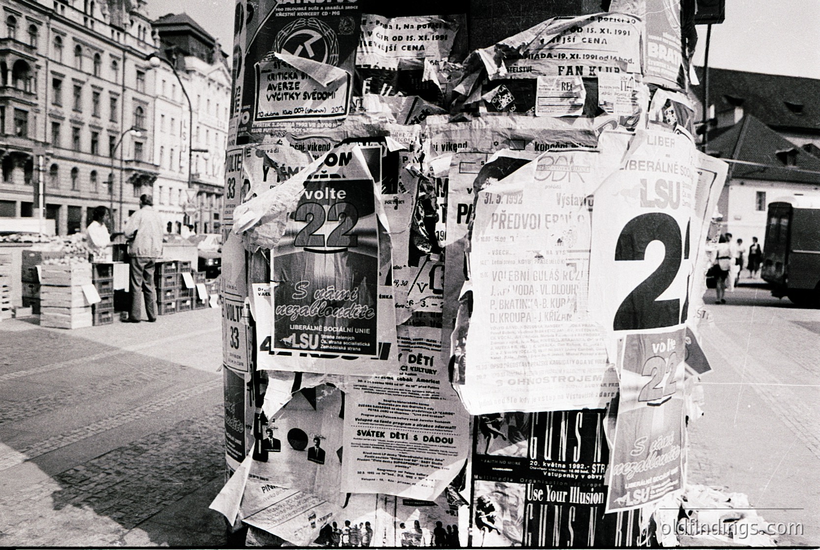 A dense cluster of vintage posters and flyers covers a lamppost in an urban setting, featuring election campaign materials (, , ). Text includes party logos, candidate numbers, and slogans in a European language. Historic European cityscape with classical architecture in background (, , ).