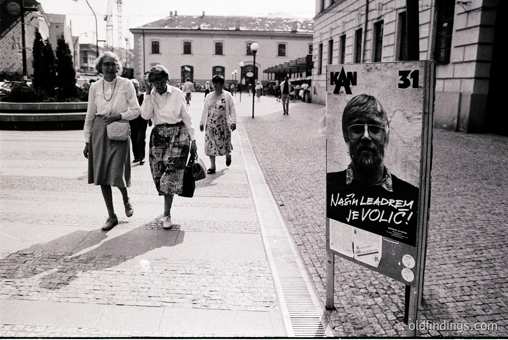 Black-and-white street scene featuring three women in 1970s-era dresses and headscarves crossing a cobblestone pedestrian crossing. Prominent poster on right reads *"Naj leadrem je Volic!"* (Slovenian: "Volic is the leader!") with a bearded man’s portrait. Urban architecture includes stone buildings and a visible "Kan 31" sign. Likely Ljubljana, Yugoslavia, 1970s.