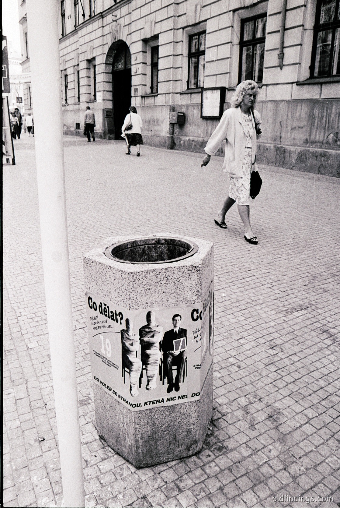 Urban trash bin with Czech/Slovak text ("Co dělat?" + silhouettes) on cobblestone street. Mid-20th century European architecture in background. Woman in 1960s-style dress walks past.