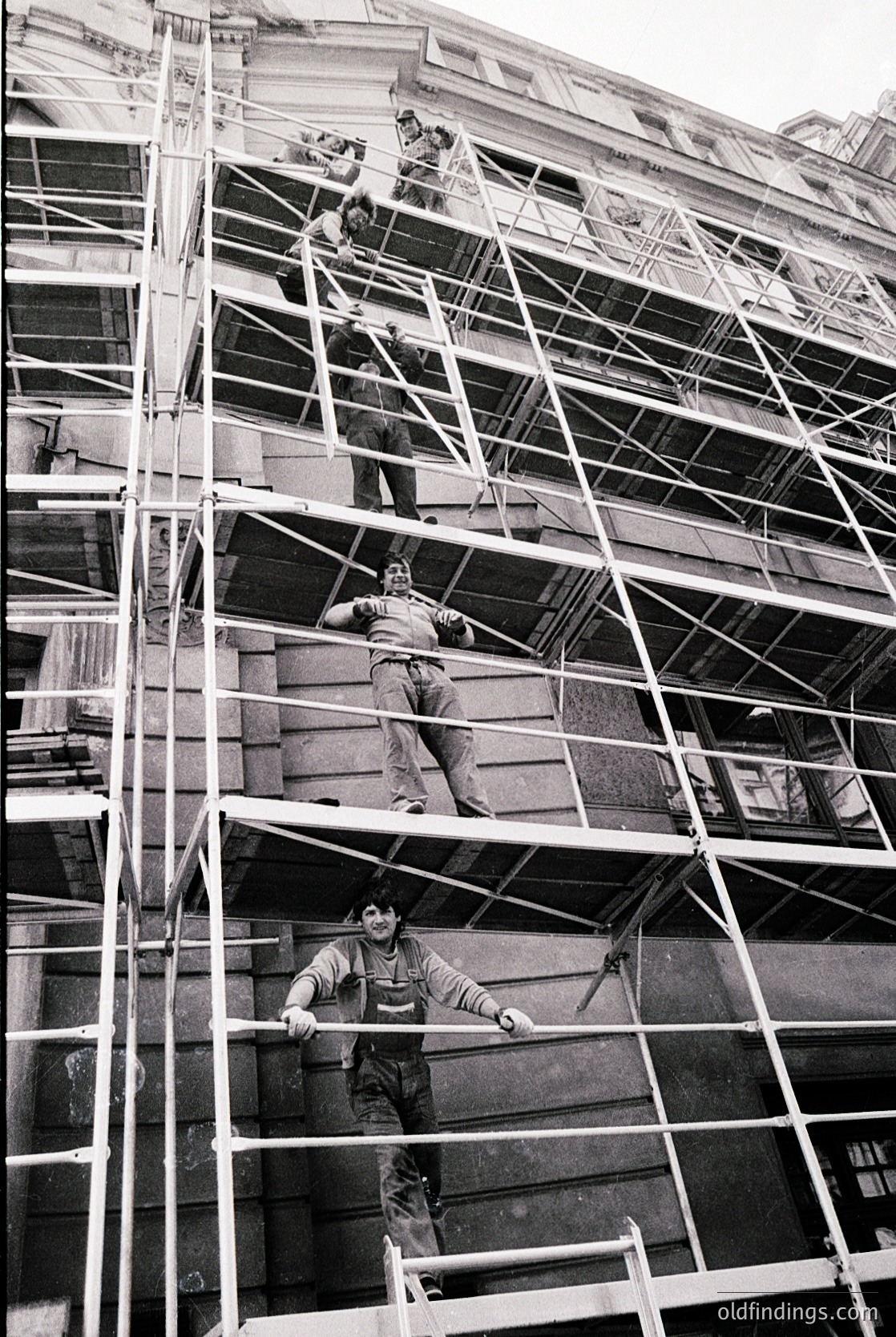 Three construction workers in harnesses ascend multi-story scaffolding on a mid-20th-century urban building. Heavy-duty metal scaffolding frames the facade, suggesting renovation or maintenance. Workers wear long-sleeve shirts, pants, and safety gear typical of the era.