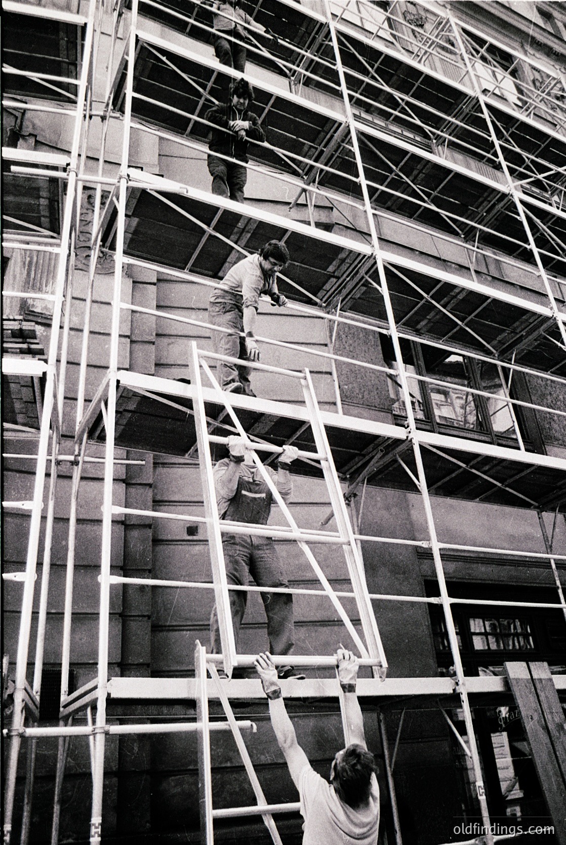 Mid-20th-century construction crew ascending metal scaffolding on a high-rise facade. Workers in casual attire (short-sleeved shirts, trousers) demonstrate mid-century labor practices. Urban setting with visible residential windows and balconies.