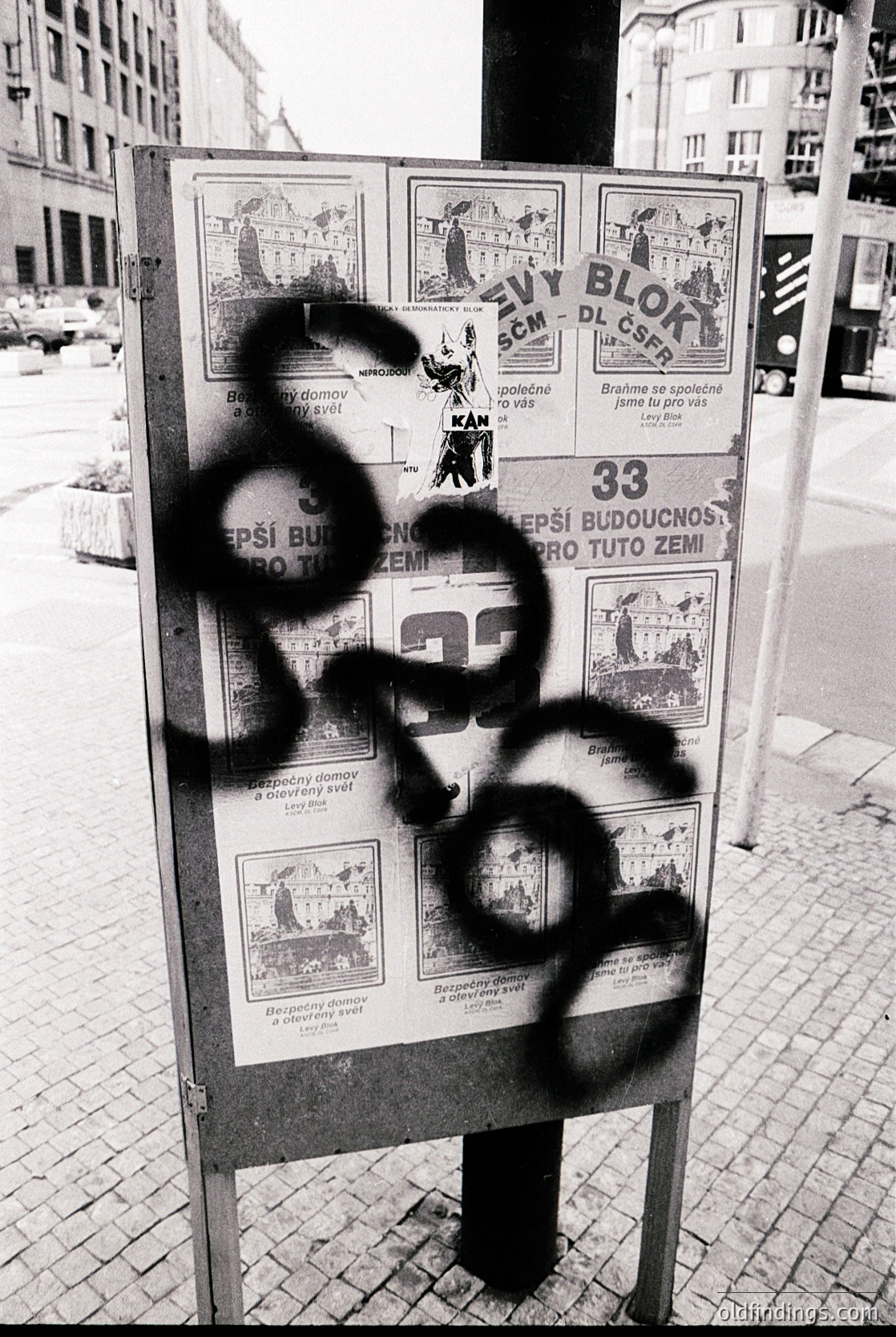 Vintage public housing campaign posters from the 1960s–70s, featuring illustrations of families and housing blocks in Czech/Slovak. Graffiti obscures one poster’s text. Urban setting with brick pavement and mid-century architecture in background.