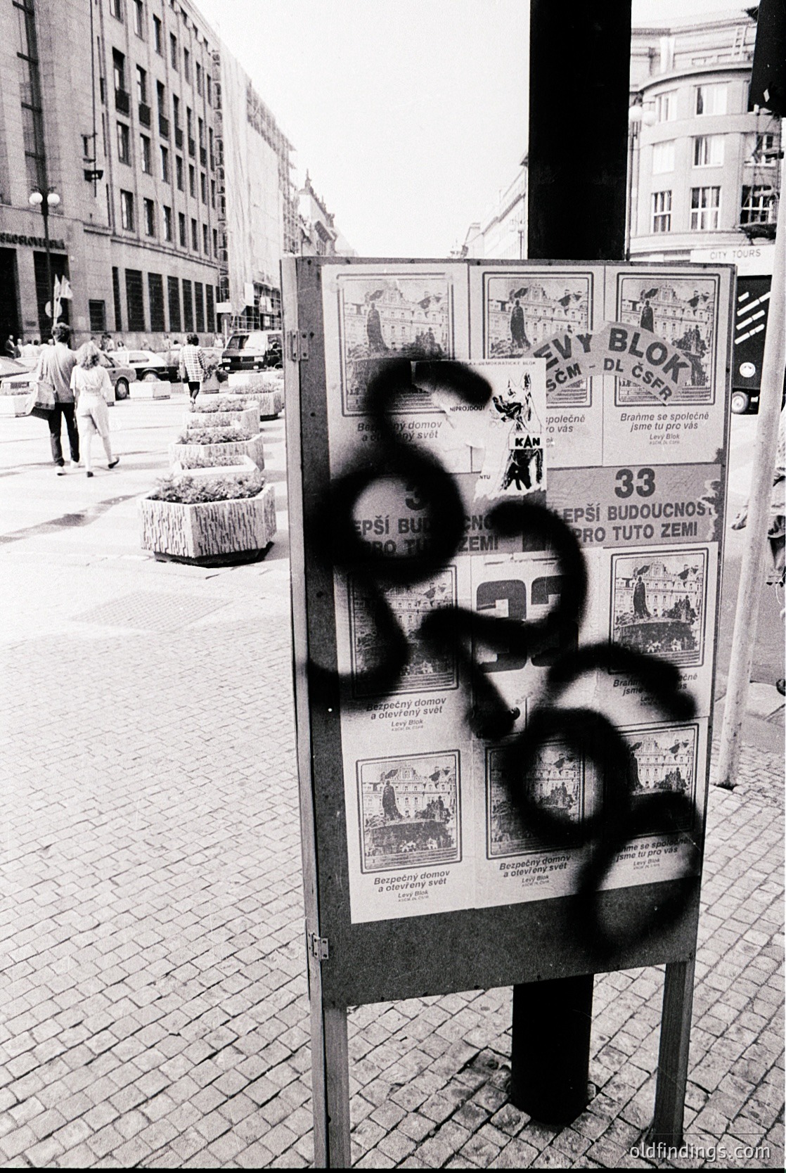 Vintage urban poster board in Eastern Bloc-style city, featuring graffiti "68" over a 1968 housing campaign ad. Buildings exhibit Soviet-era architecture with large windows and concrete facades. Pedestrians and parked cars suggest a mid-20th-century European street scene.