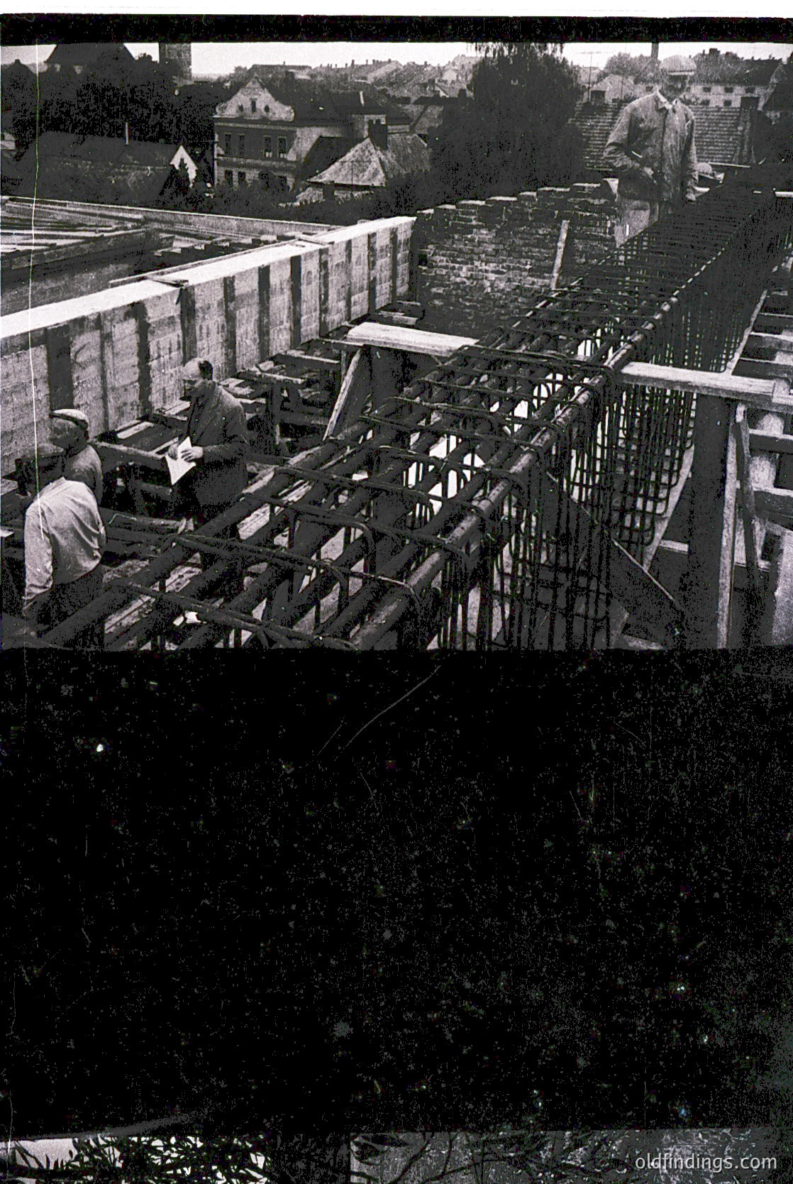 Mid-20th century construction site featuring reinforced concrete framing. Workers in casual attire (short sleeves, caps) assemble steel rebar scaffolding. Urban backdrop includes residential buildings and railway tracks. Likely Eastern European, 1950s-1960s.