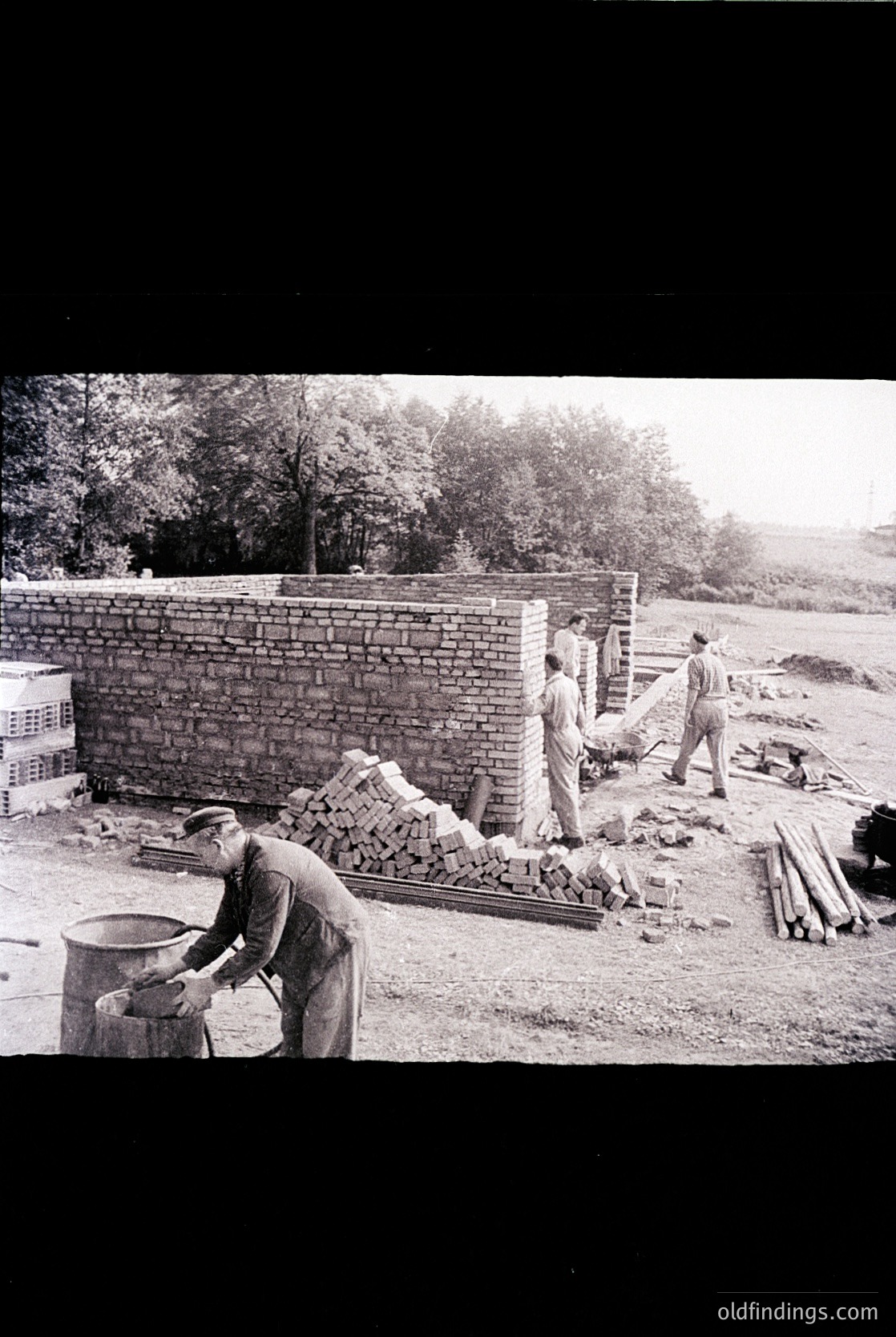 Mid-20th century bricklaying crew constructing a wall, likely . Workers in rolled-up sleeves, using bricks and mortar; stacked lumber and bricks on-site. Rural or industrial setting with trees in background.