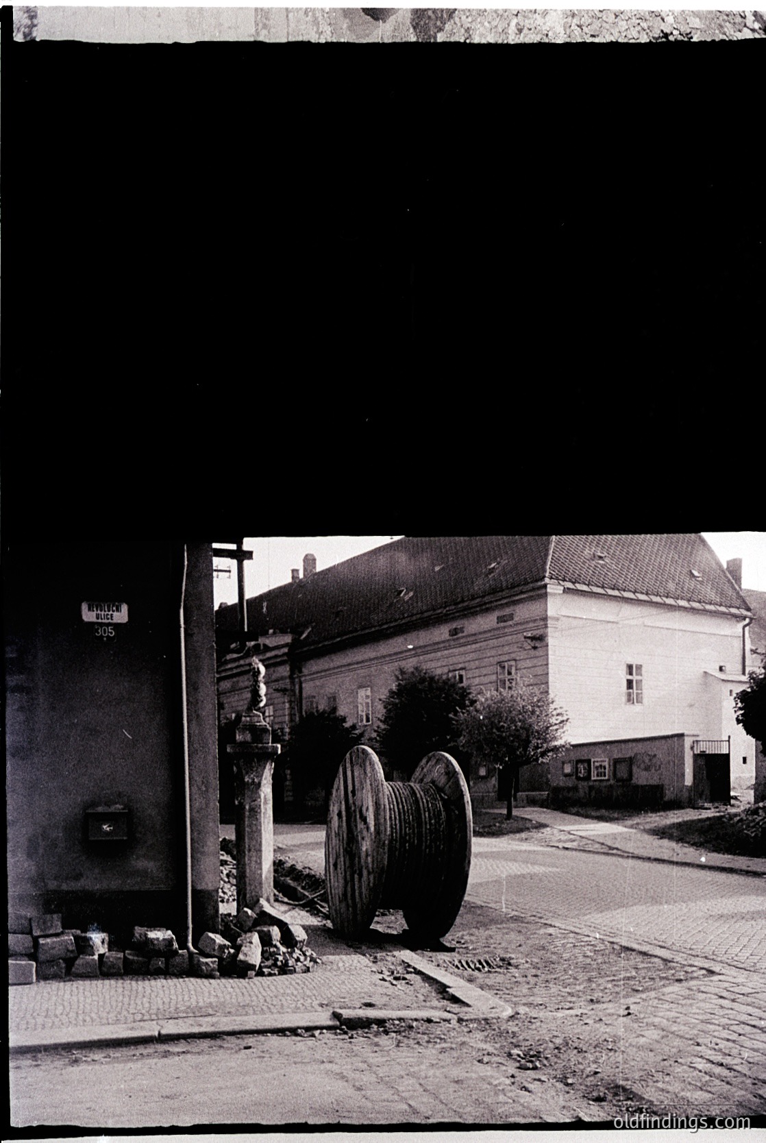 Vintage street scene featuring a large wooden water wheel beside a cobblestone sidewalk. Adjacent brick building with a fireplug and utility box. Mid-20th century European urban architecture.