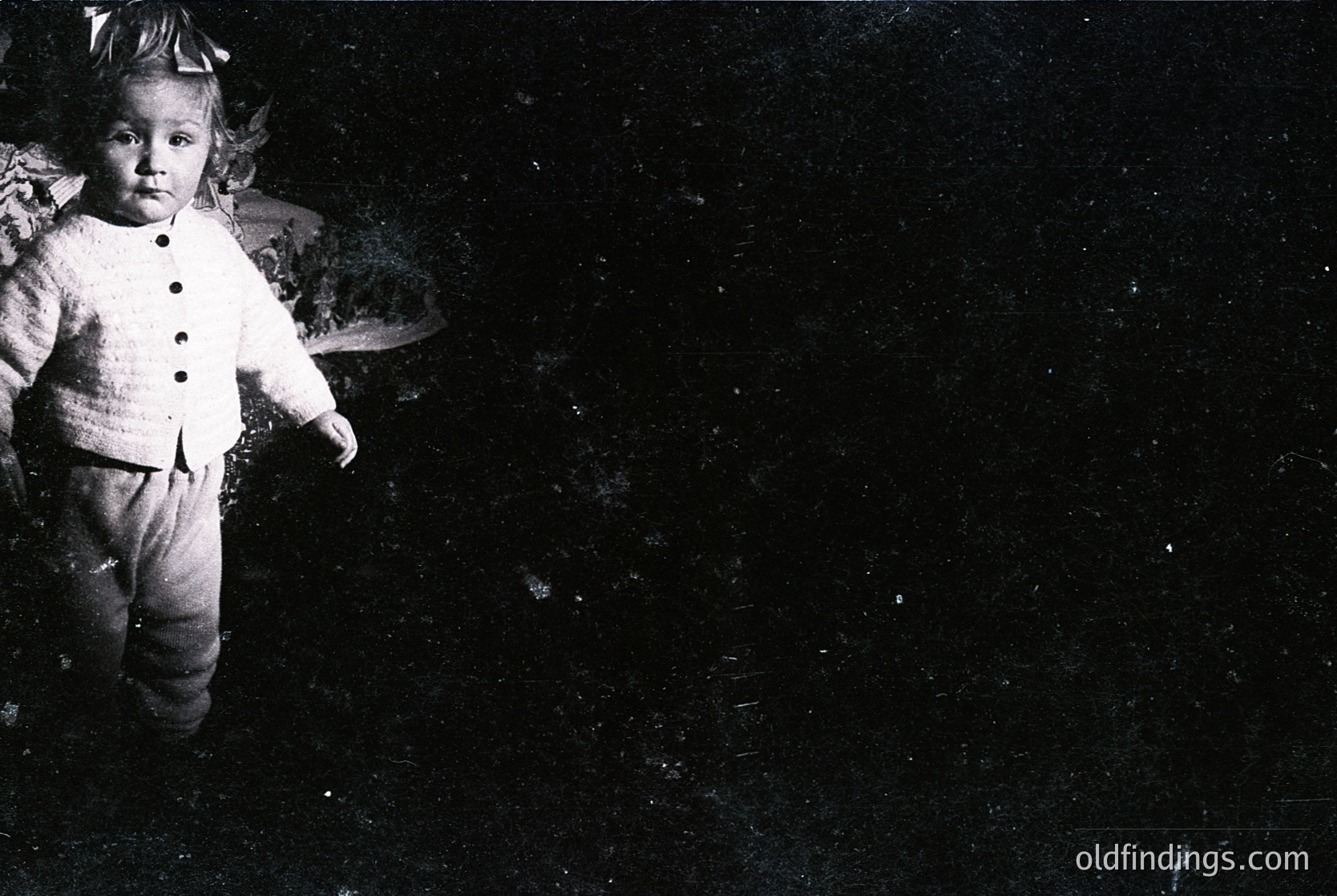 Black-and-white portrait of a young child in early 20th-century attire—buttoned sweater, trousers, and hat—posing indoors against a dark, textured background. Likely mid-1900s, possibly for studio portraiture.