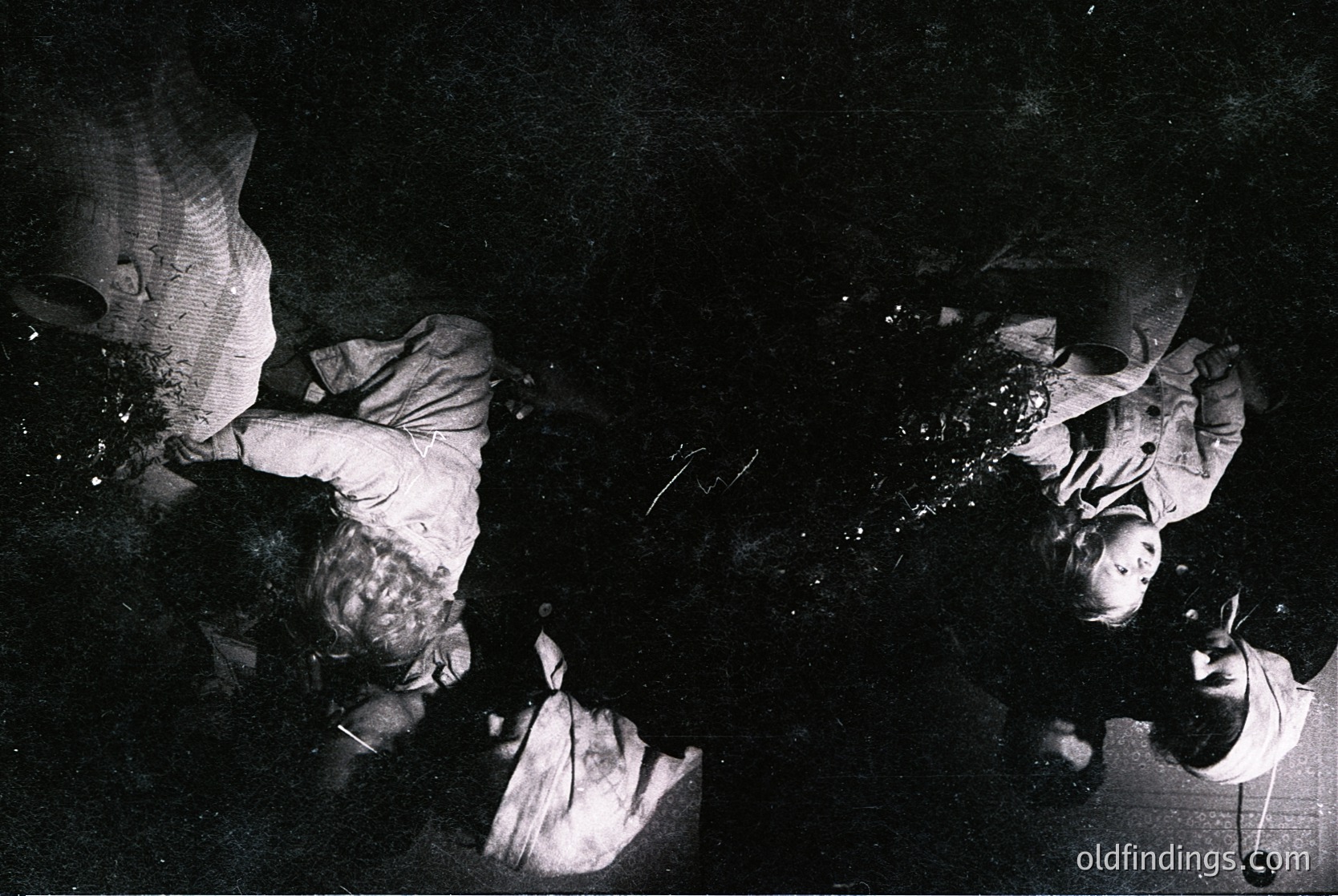 Black-and-white photo of two individuals engaged in textile work under dim lighting, likely a loom. Cloth appears partially woven, with visible threads and a partially completed fabric. Setting suggests a workshop or home-based crafting environment, indicative of traditional textile production. Style and lighting suggest mid-20th century, possibly .