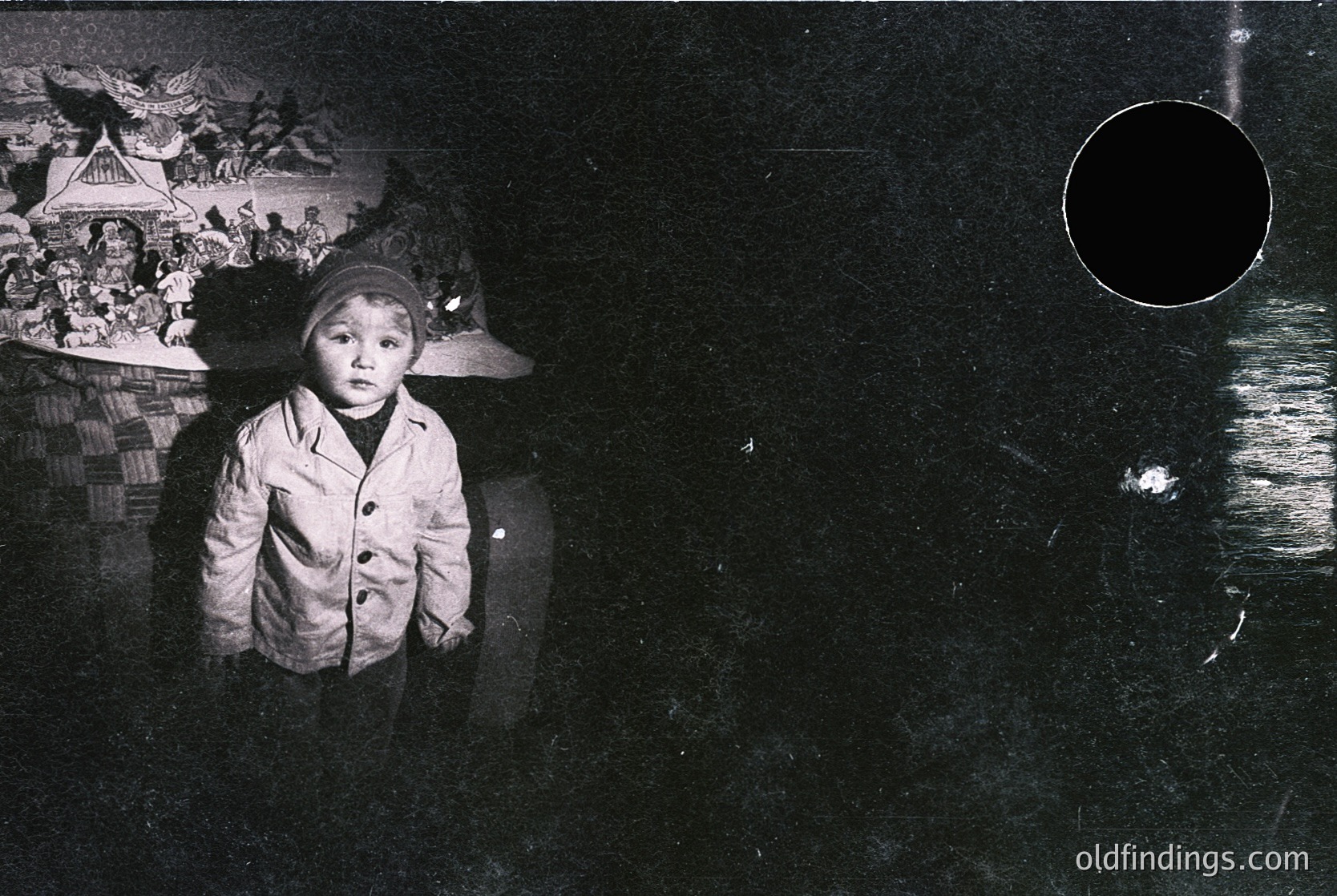 Black-and-white portrait of a young child in a heavy winter coat and knitted cap, standing indoors near a dark, reflective surface. Behind them, a mural depicts a rural scene with traditional architecture and people. Likely mid-20th century (1940s–1960s) European or Soviet-era aesthetic.