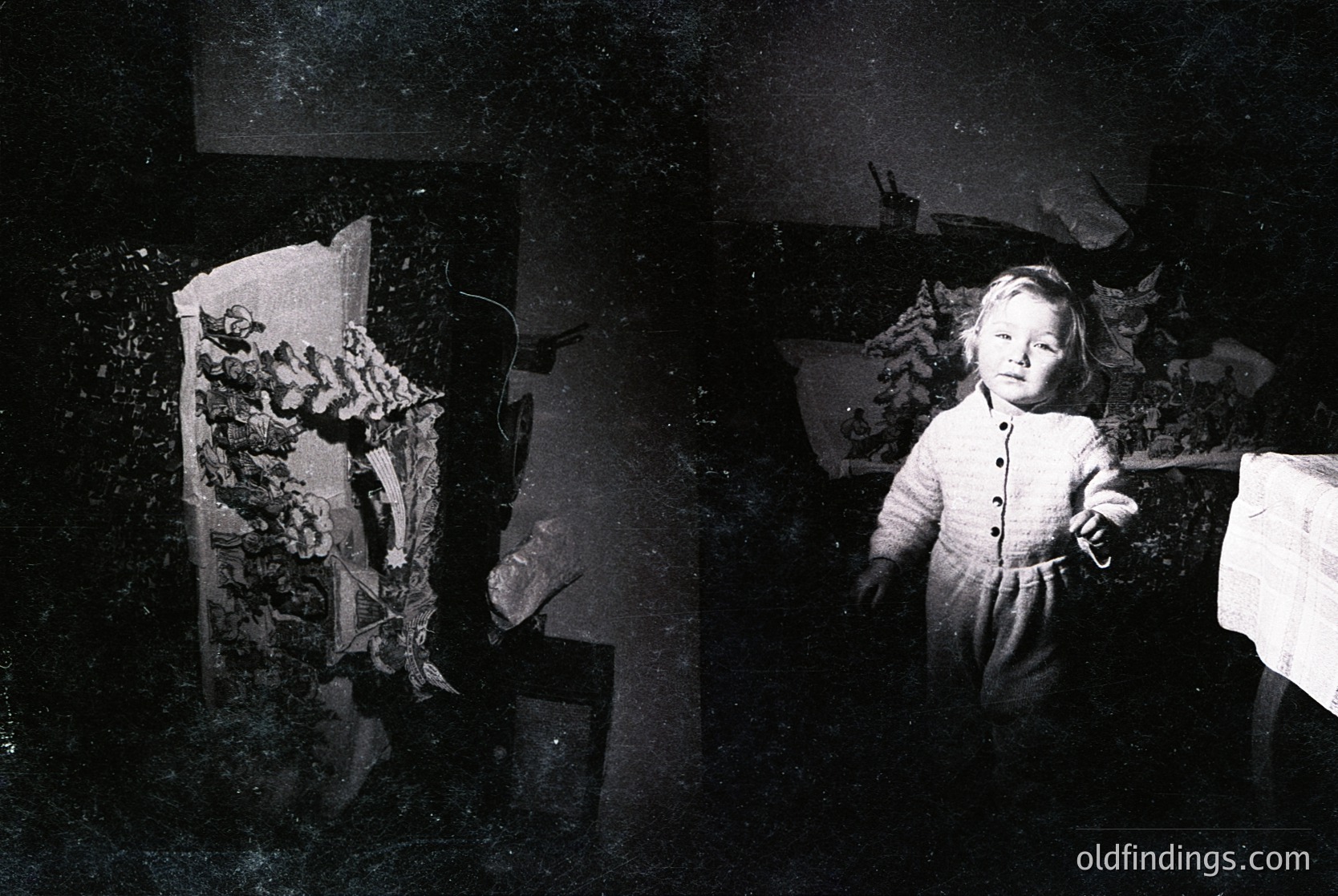 Vintage black-and-white photo of a young child in a floral-patterned dress holding a small object, likely a toy or key. Dimly lit room with visible furniture and a curtain. Mid-20th century domestic setting.