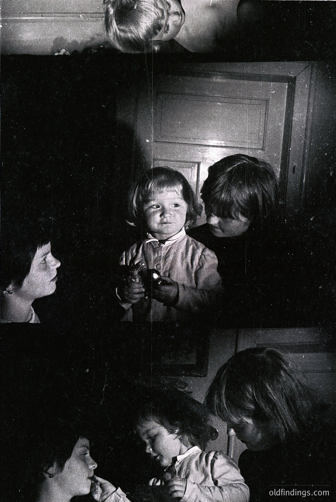 Vintage black-and-white photo of a family moment: a woman holding a child in a doorway, another child peering over her shoulder, and a third child seated below. The setting appears domestic, likely mid-20th century. The woman’s attire and lighting suggest a candid, intimate snapshot.