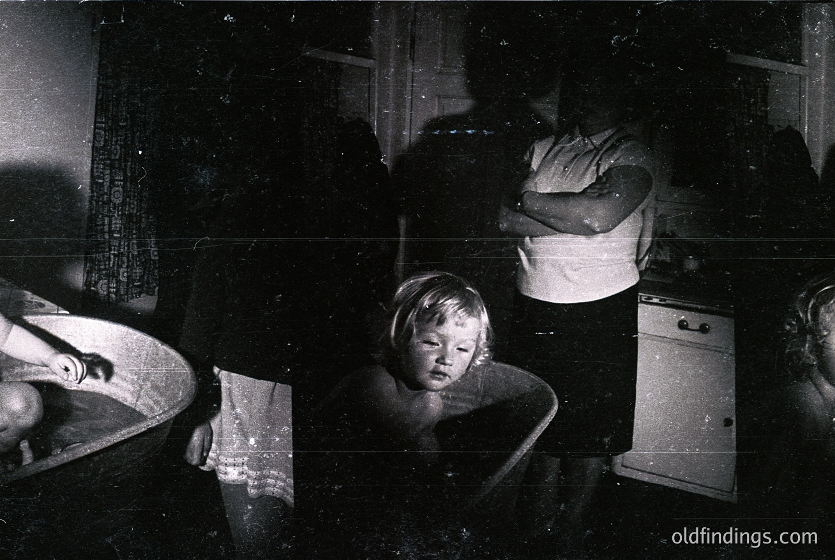 Vintage black-and-white photo capturing a dimly lit kitchen scene, likely mid-20th century. A woman kneels beside a child seated on a stool, both facing a sink with a visible faucet. A vintage stove and wooden cabinetry frame the composition. Warm, intimate domestic moment.