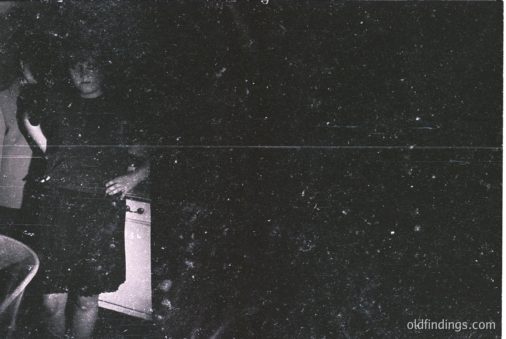 Vintage black-and-white photo of a person’s lower body seated on a wooden chair, holding a small, upright object resembling a vintage camera or projector. Grainy texture suggests early 20th-century film photography. Indoor setting with dark, speckled background.