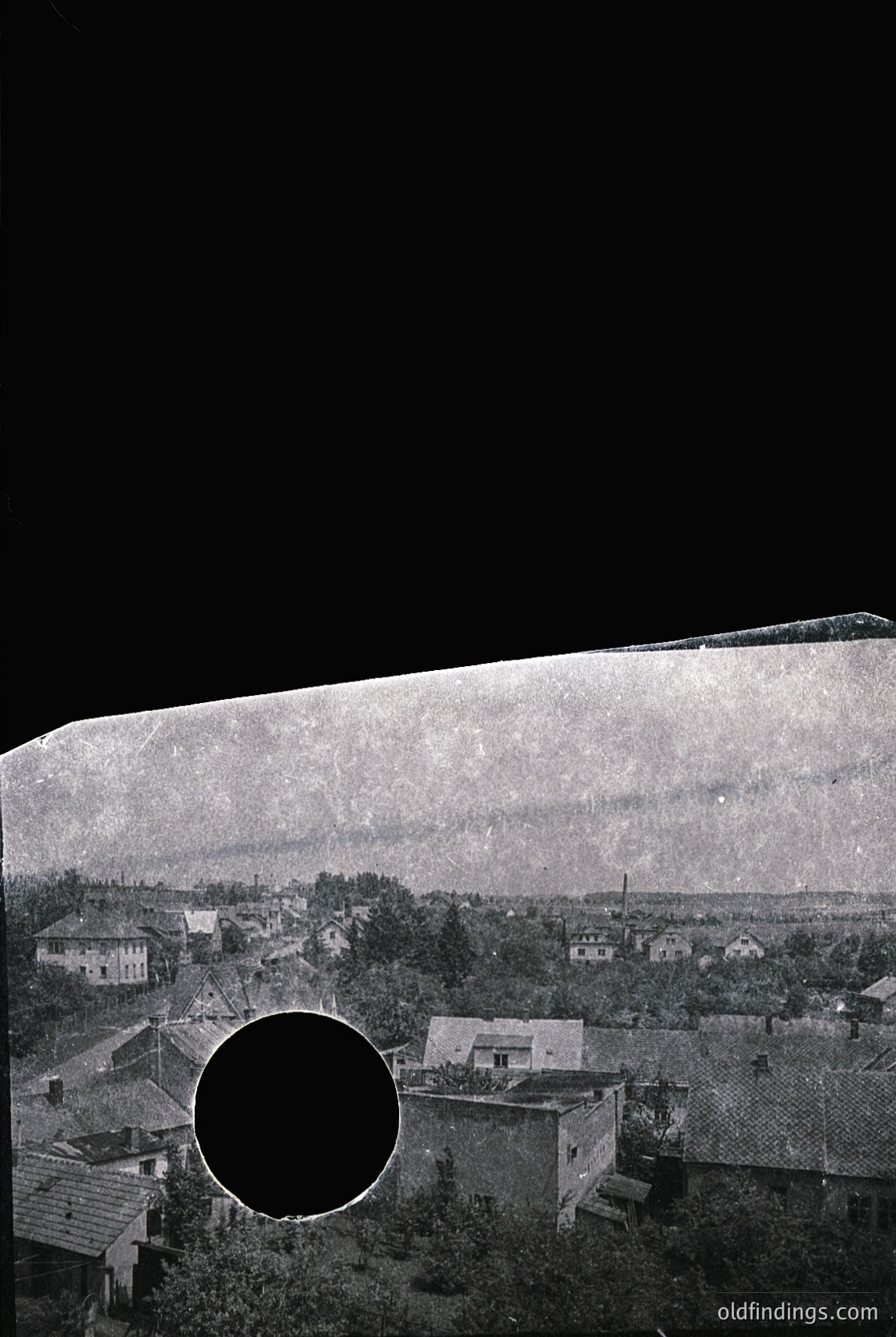 Vintage viewfinder photograph showing a rural settlement with low-slung brick houses and dense greenery. Circular aperture reveals a framed perspective, likely from a 19th–early 20th century camera.