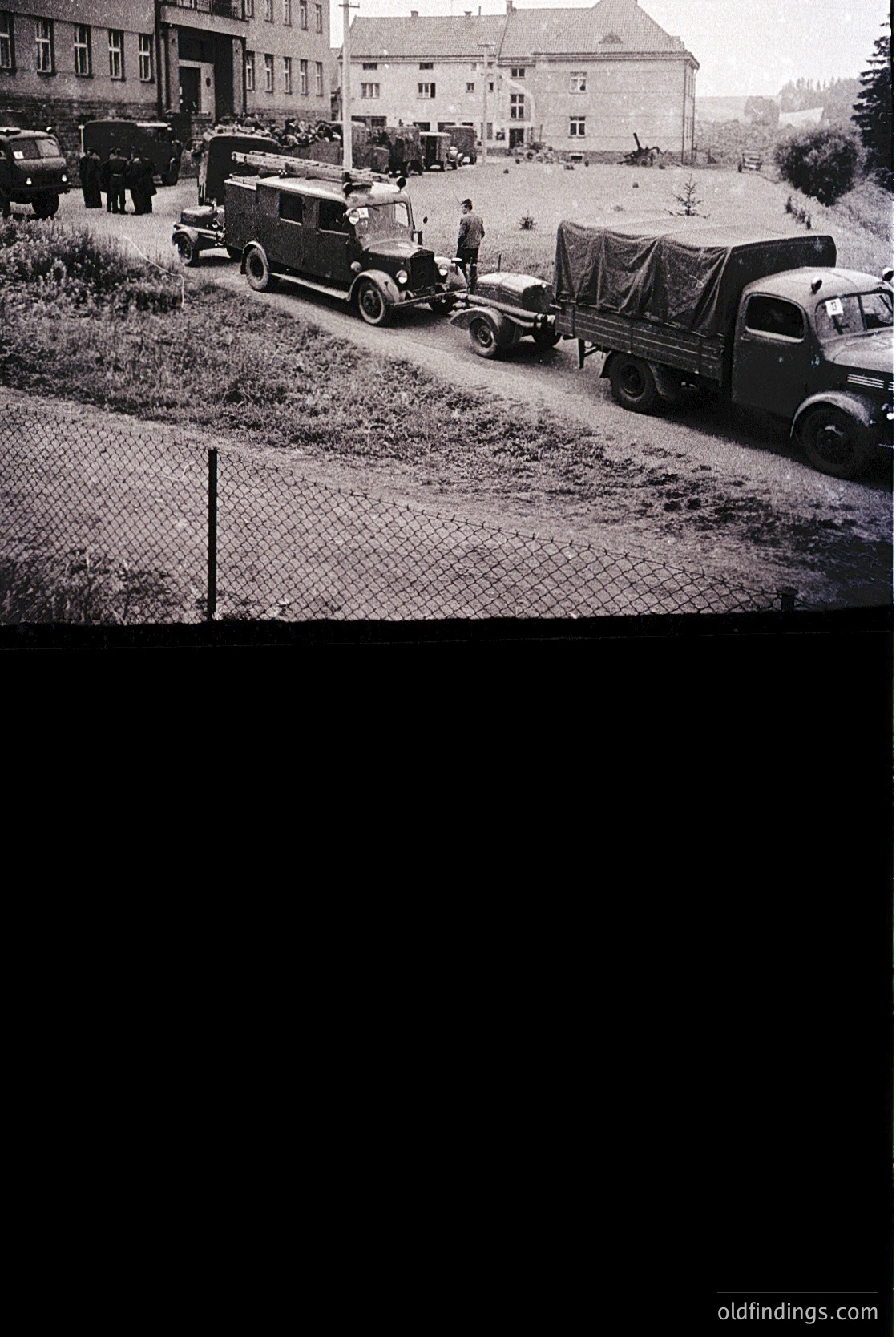 Vintage black-and-white photo of a rural roadside scene with vintage military trucks and a jeep, likely mid-20th century. Open cargo beds carry supplies or equipment, and a man in uniform stands near a truck. Brick buildings and a chain-link fence frame the background.